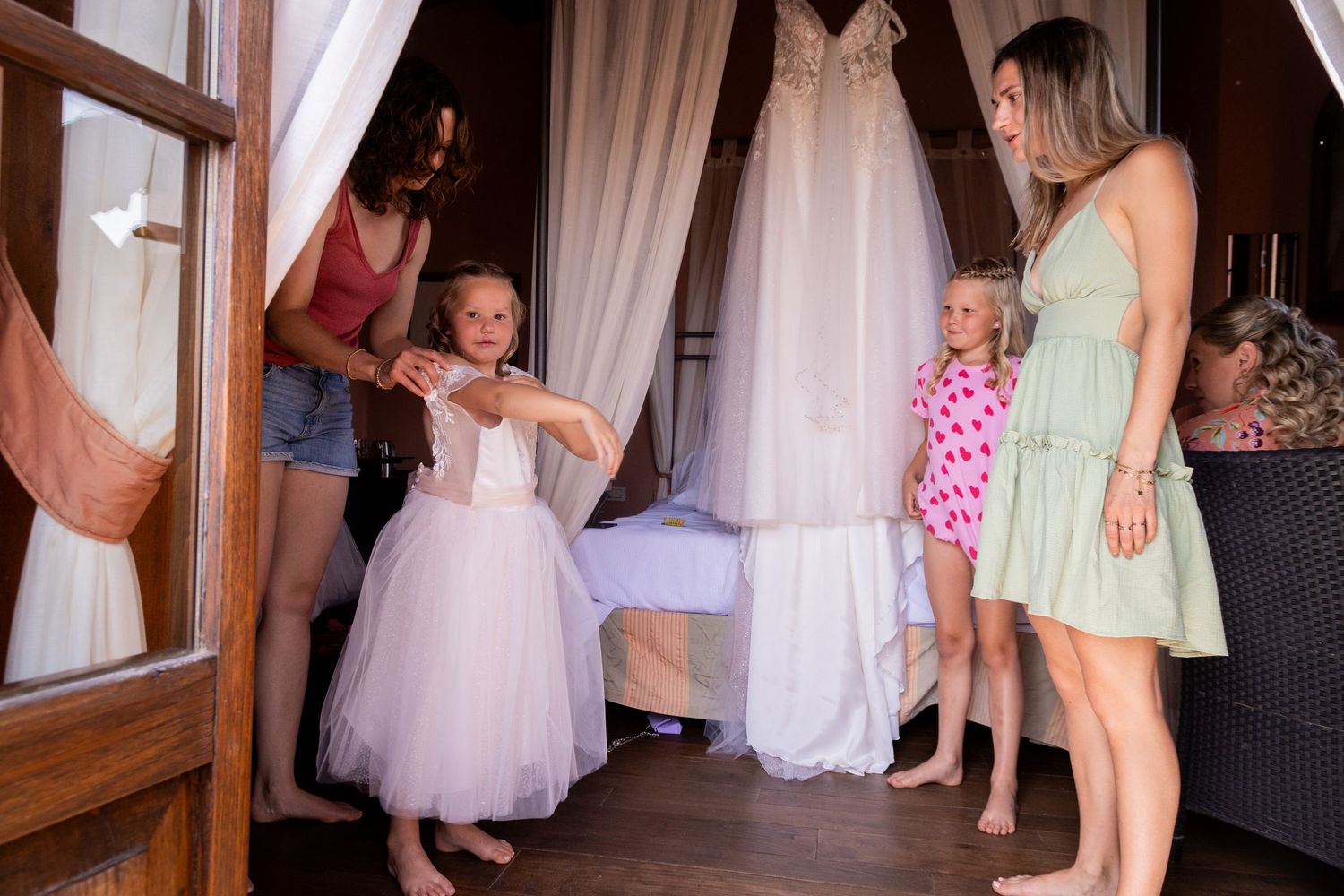 Group gathered around a white wedding dress hanging in a room with flowy curtains and soft natural lighting.