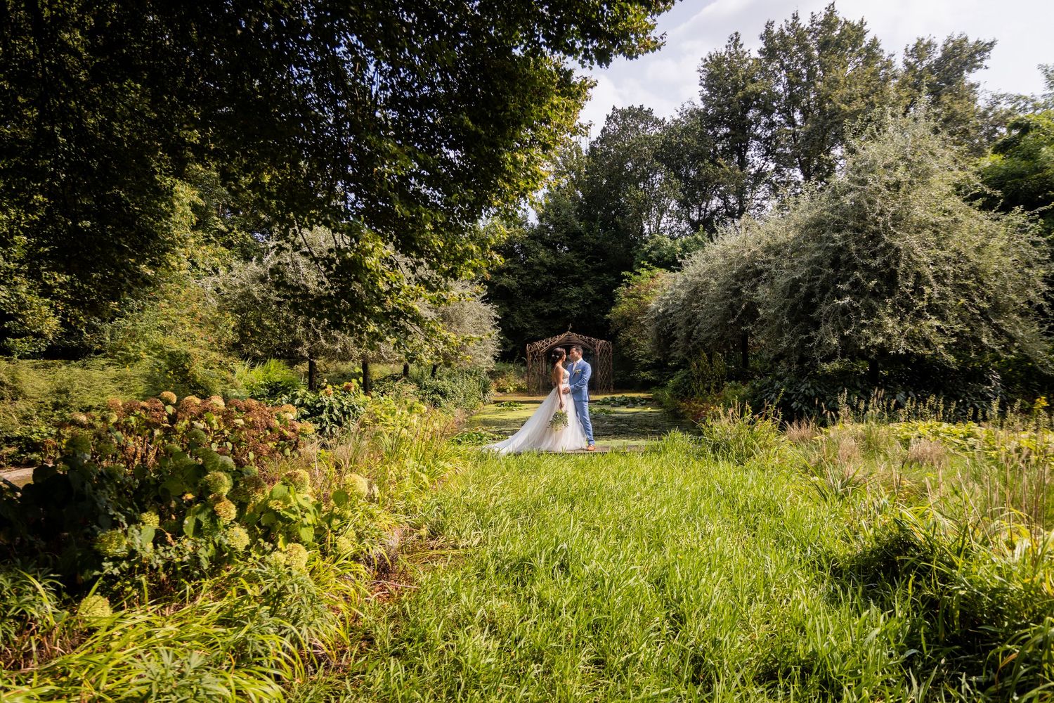A couple stands at a distance in a lush green garden path surrounded by vibrant summer foliage and trees.
