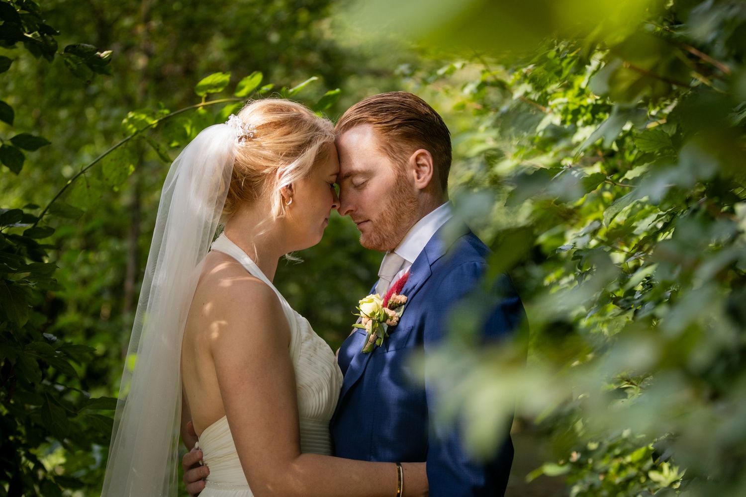 Intimate wedding portrait through green foliage with soft natural lighting.
