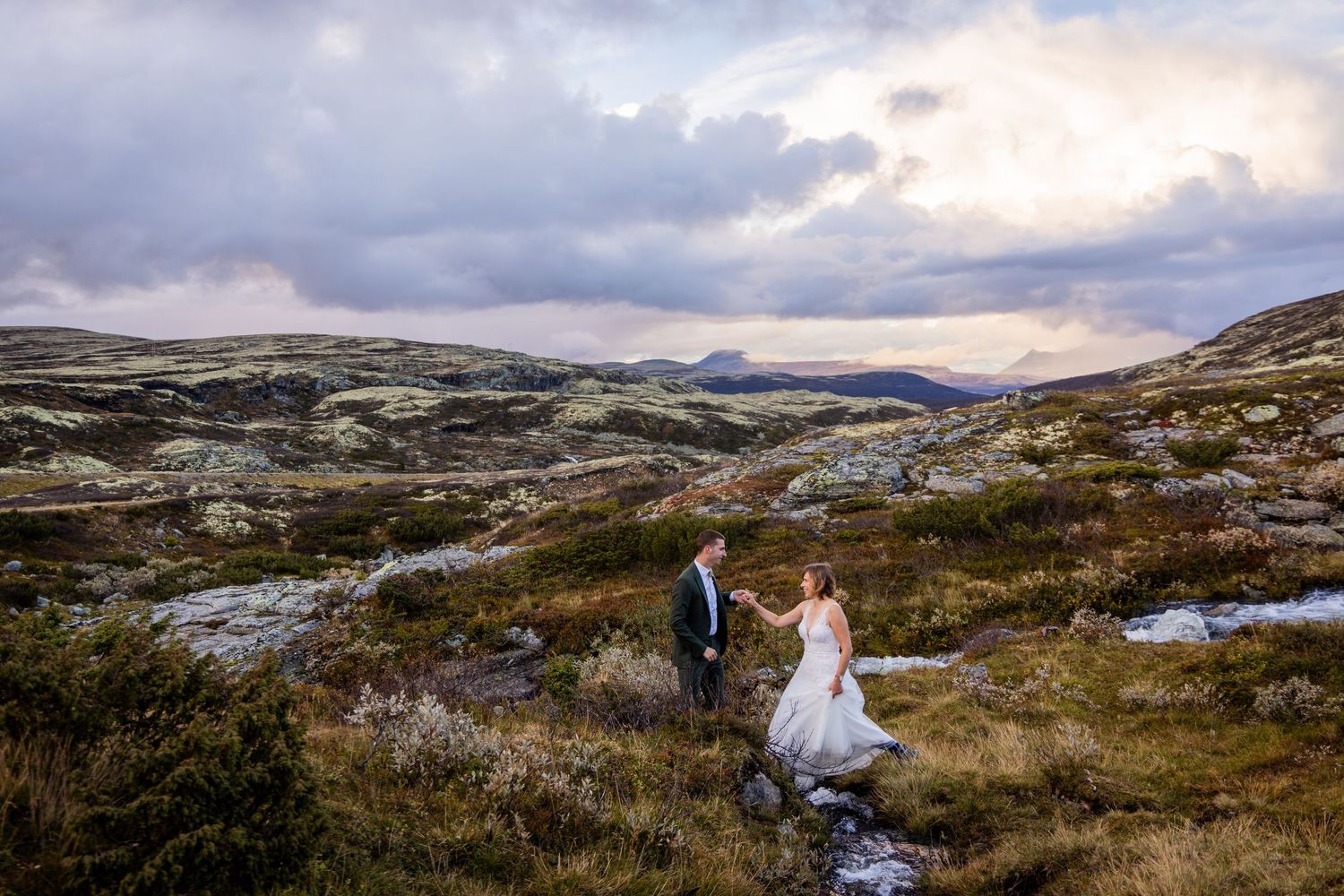 Two figures in wedding attire stand together on a rugged mountain landscape under dramatic cloudy skies in Norway.