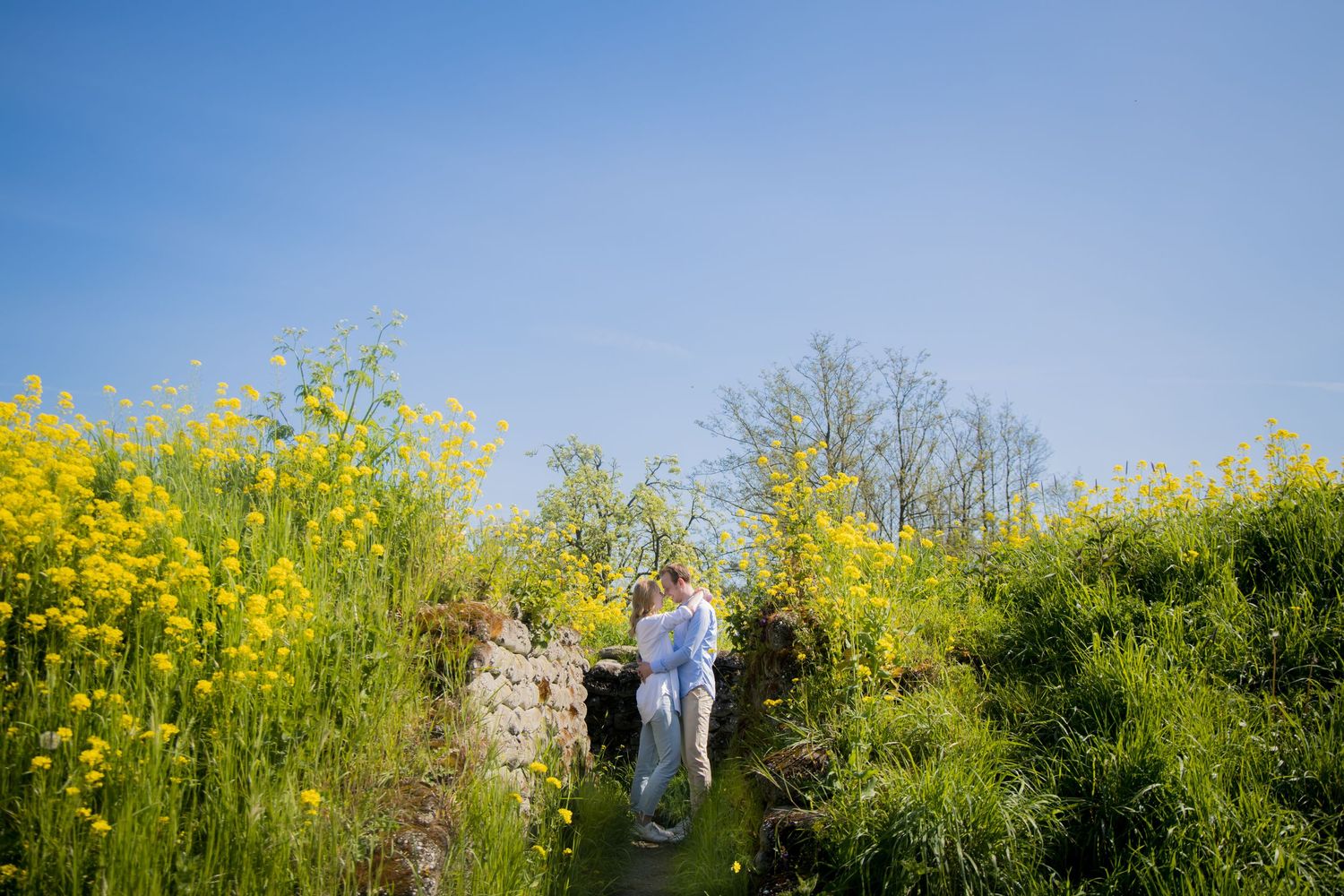 A couple stands atop stone steps surrounded by yellow wildflowers against a bright blue sky.