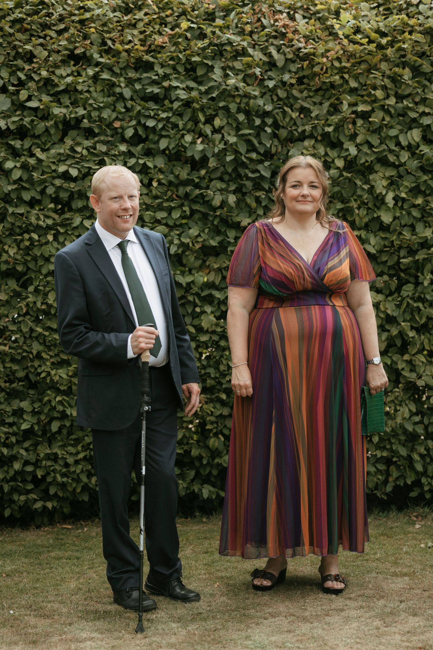 Couple in formal attire stand before green hedge at outdoor wedding reception.