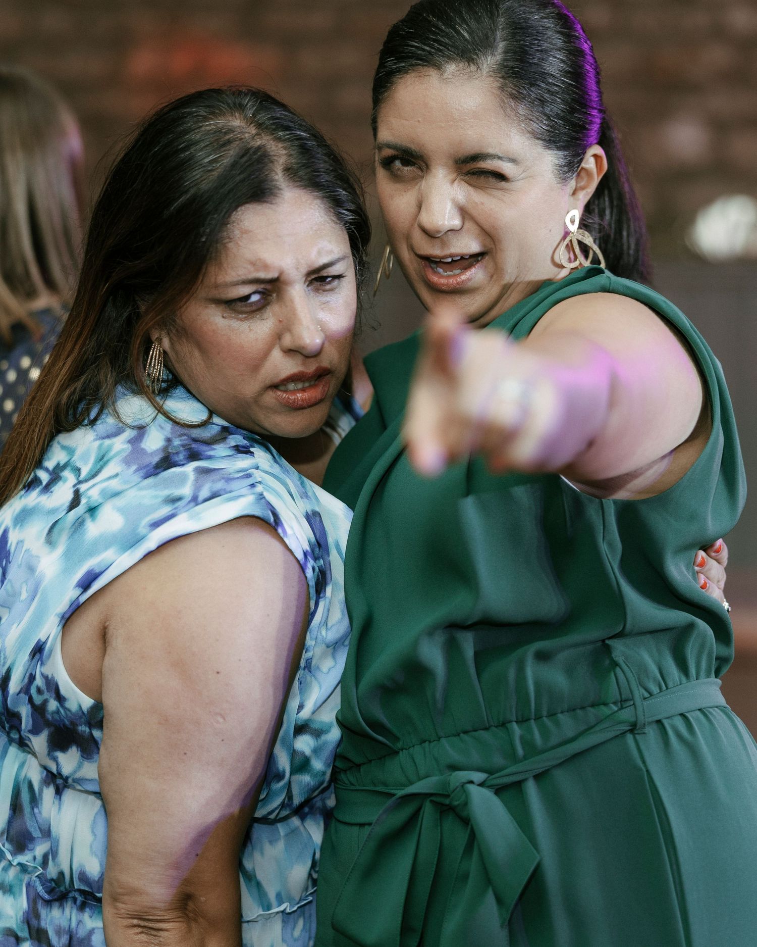 Two women in conversation, one in green dress pointing, other in blue floral top, outdoors event setting.