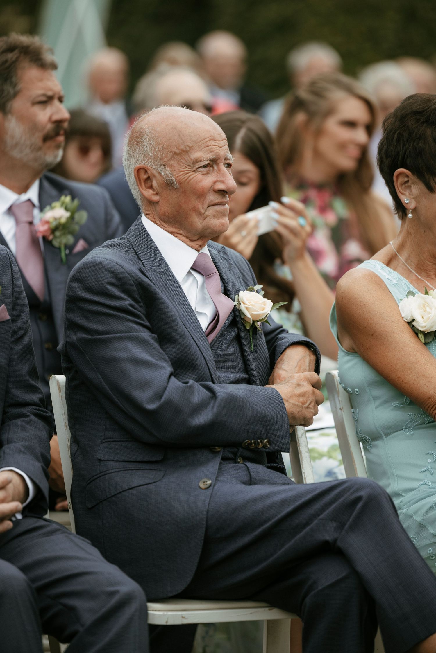 Elderly gentleman in navy suit with boutonniere seated at outdoor wedding ceremony smiling warmly.
