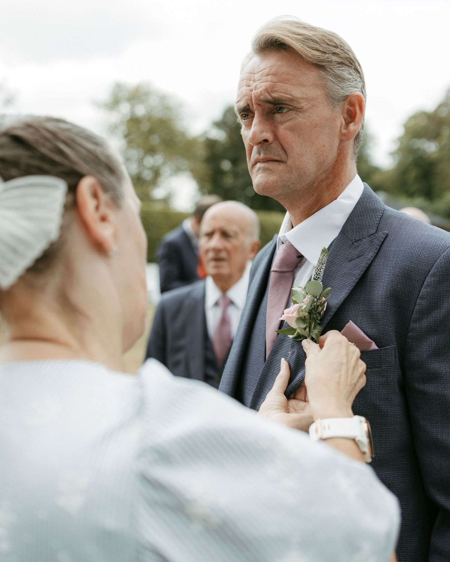 Man in navy suit having boutonniere pinned to lapel by woman at outdoor wedding celebration.