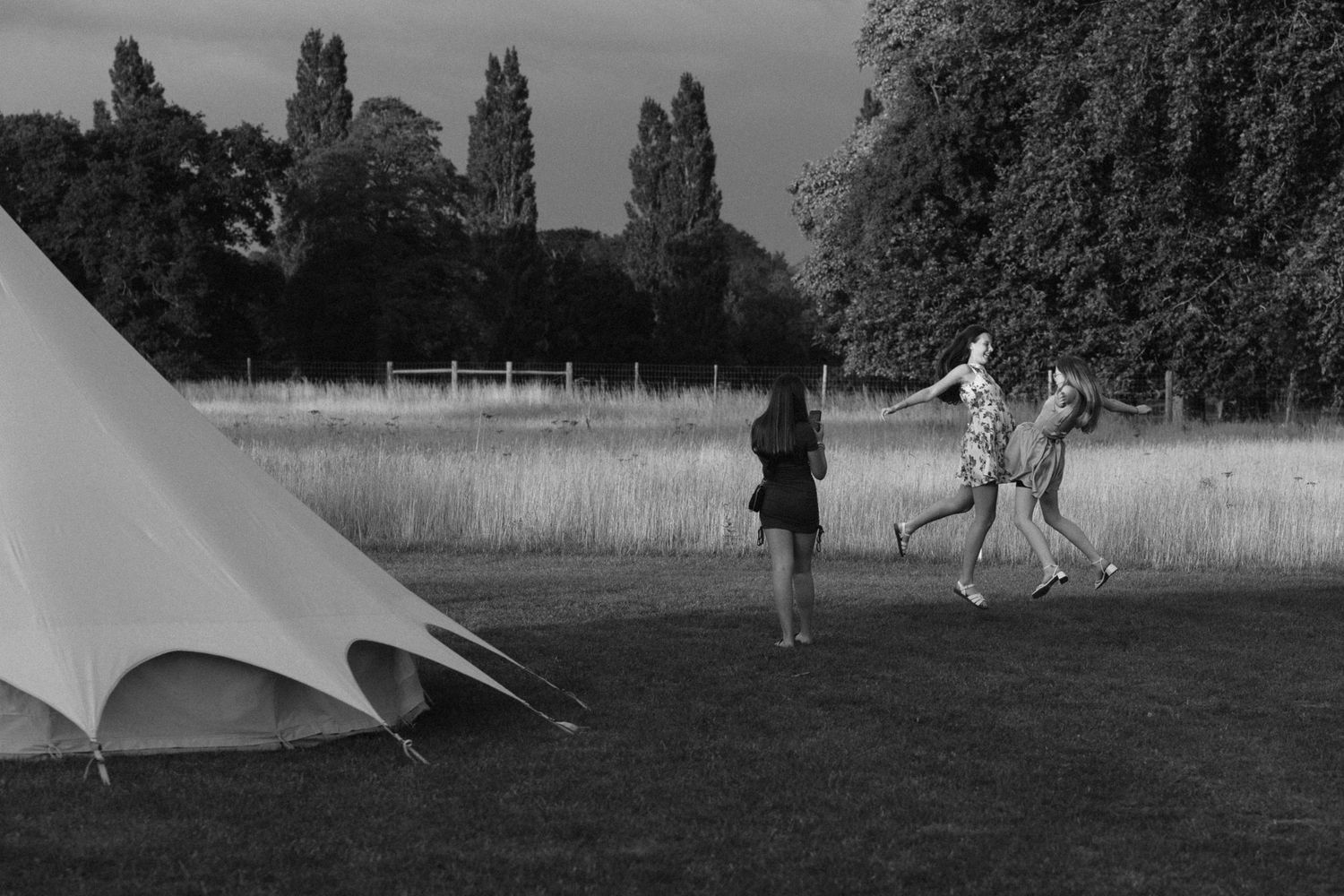Black and white photo of women playing near a teepee tent in a lush garden field with tall trees.