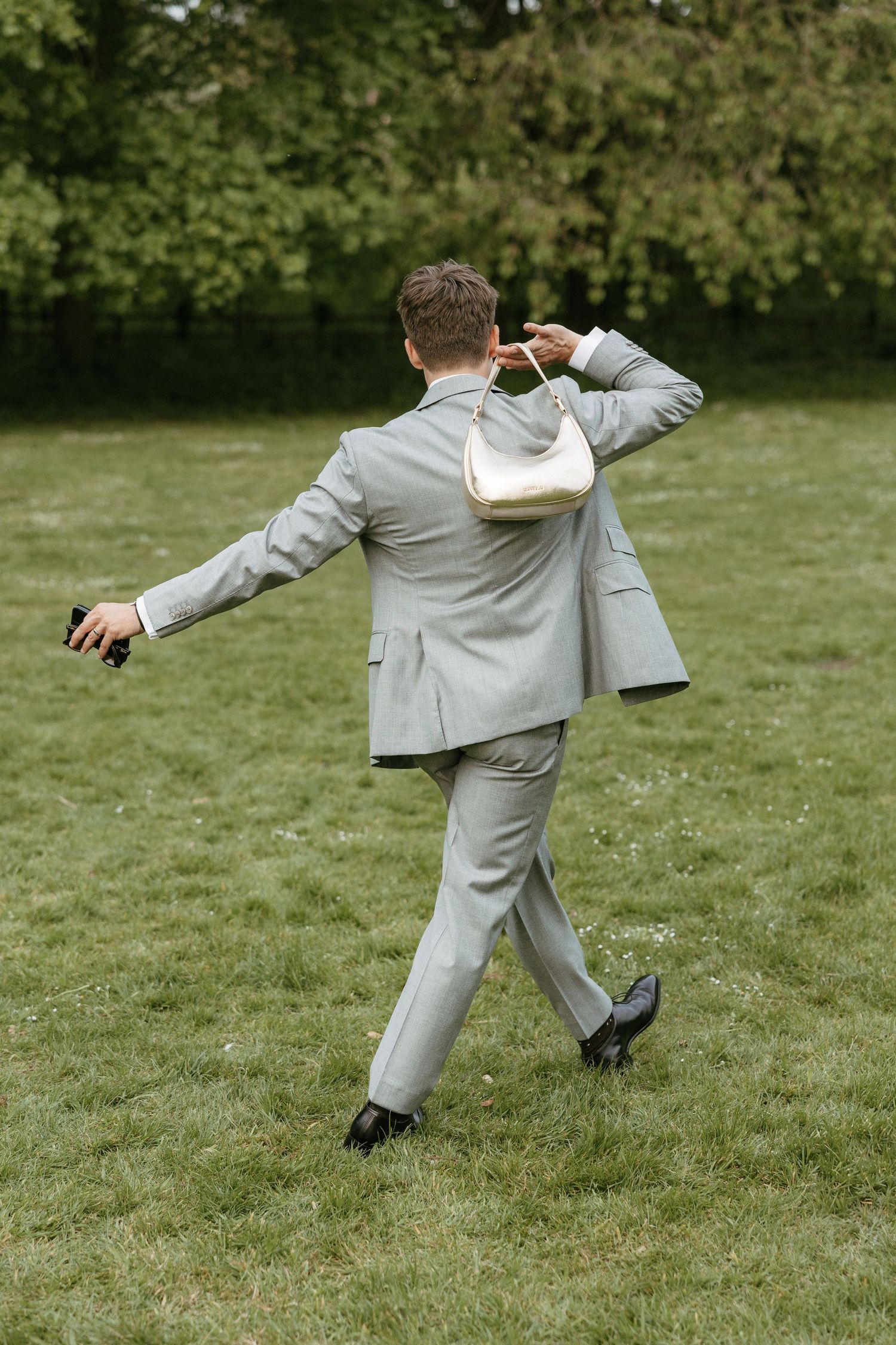 Man in light gray suit walking on grass, carrying a white handbag over his shoulder in a park setting.
