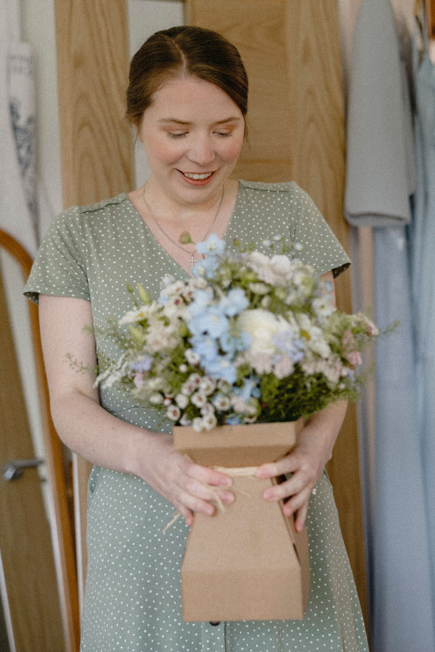 Smiling woman in sage green polka dot dress holding a pastel blue and white floral arrangement in a kraft box.