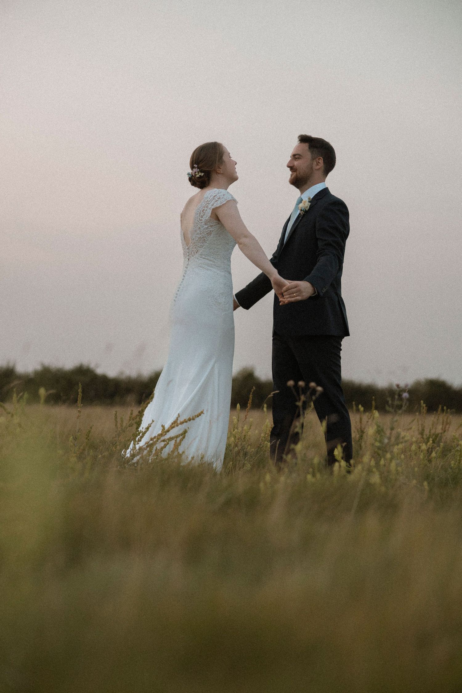 Bride and groom holding hands in a golden field at dusk, sharing a romantic moment on their wedding day.