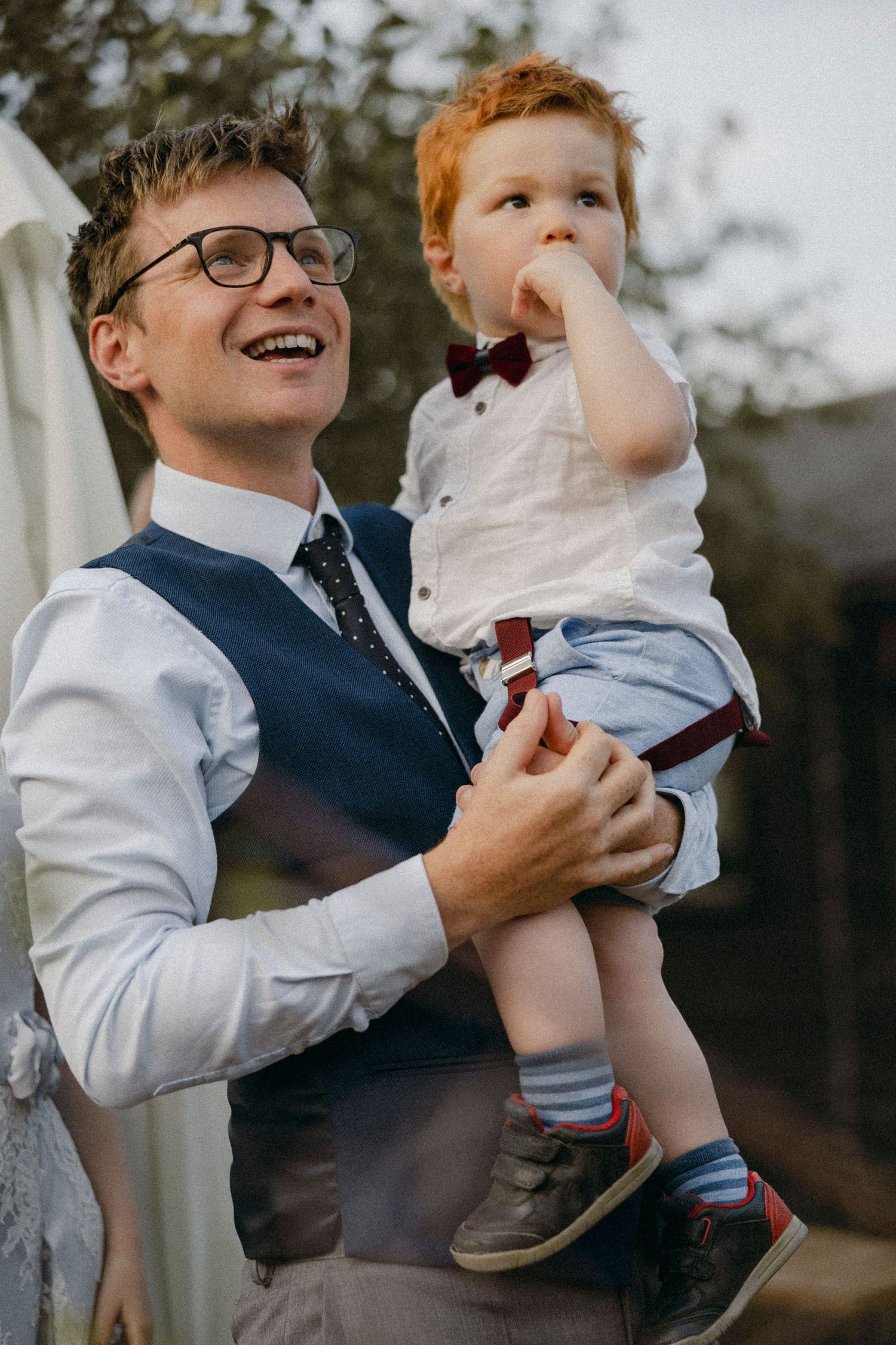 Smiling man in vest and tie holds a toddler boy in bow tie and suspenders at an outdoor event.