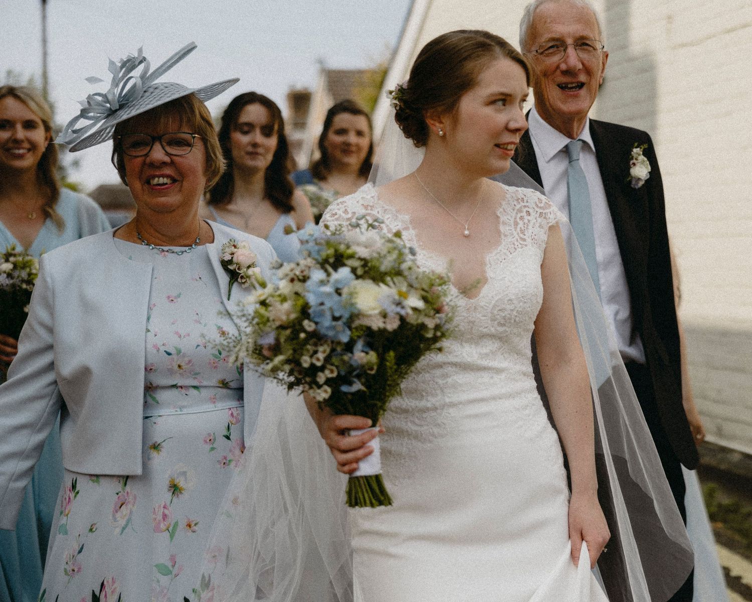Bride in white lace gown holds blue floral bouquet, walking with smiling family members at outdoor wedding ceremony.