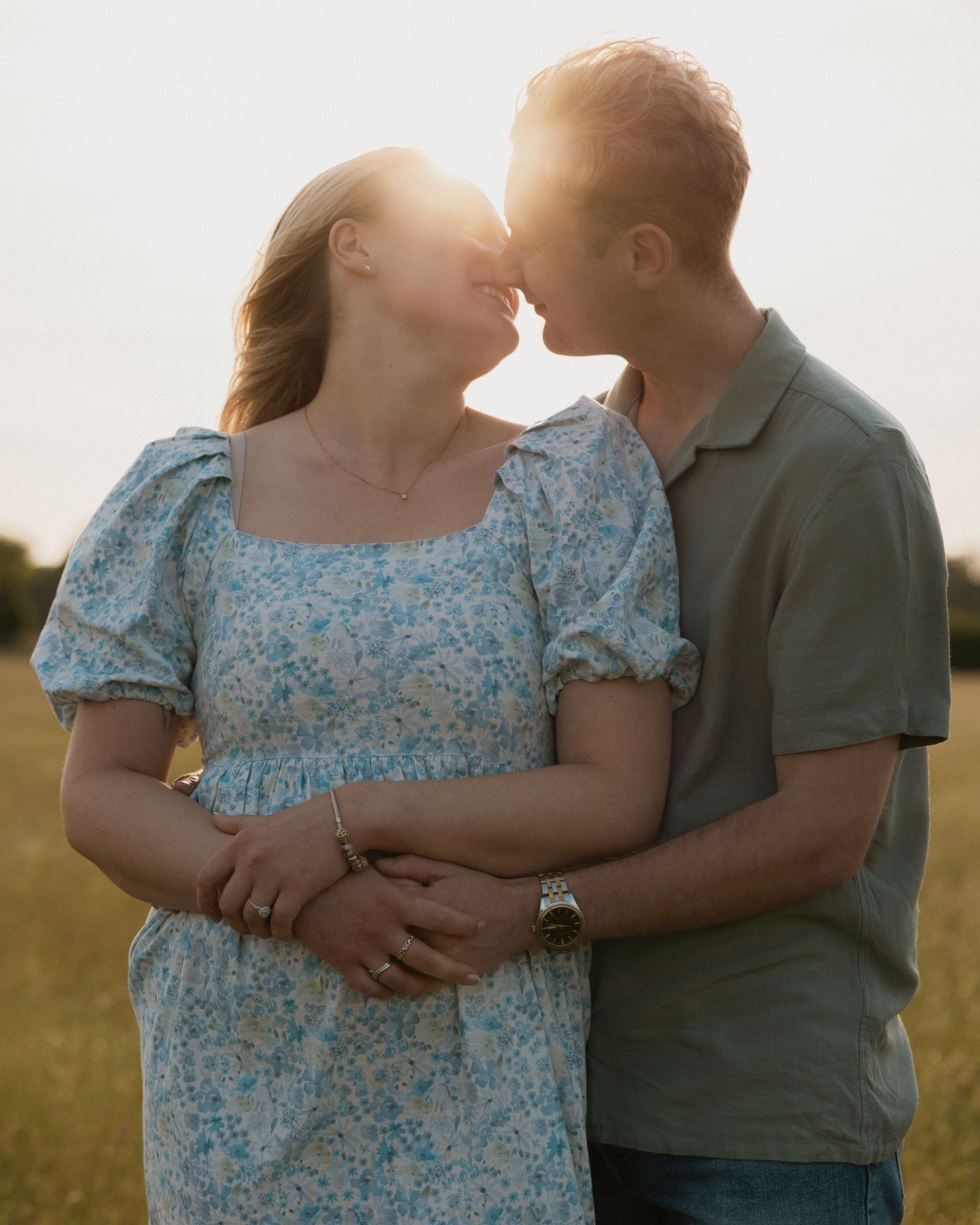 Loving couple in a golden sunset field, woman in floral dress embraced by man in green polo shirt.