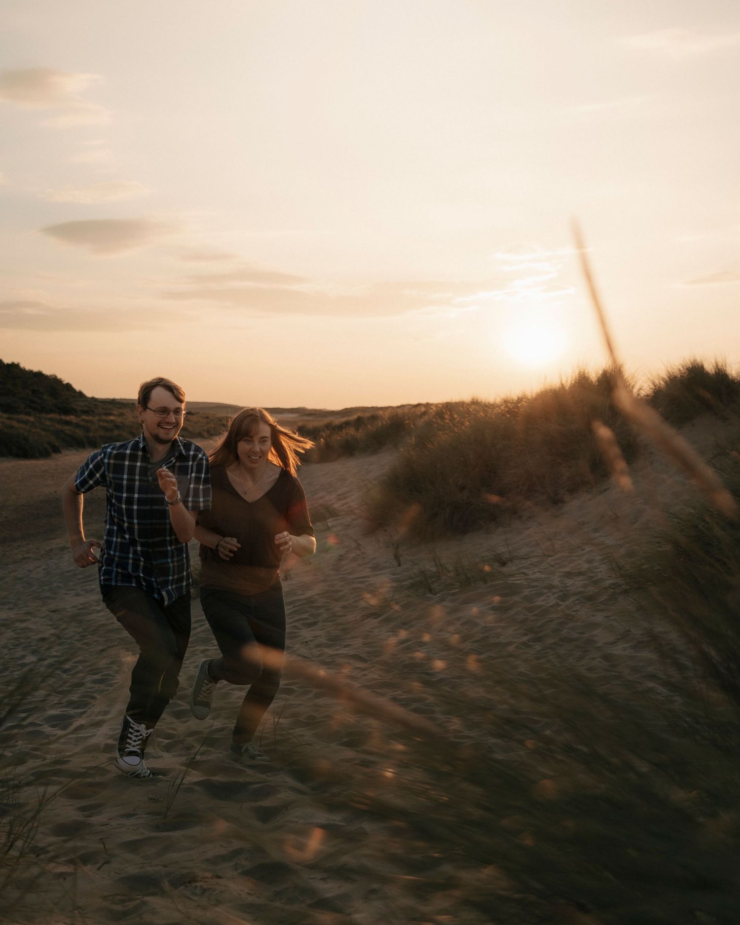 Couple laughing and running together through sandy dunes at sunset.