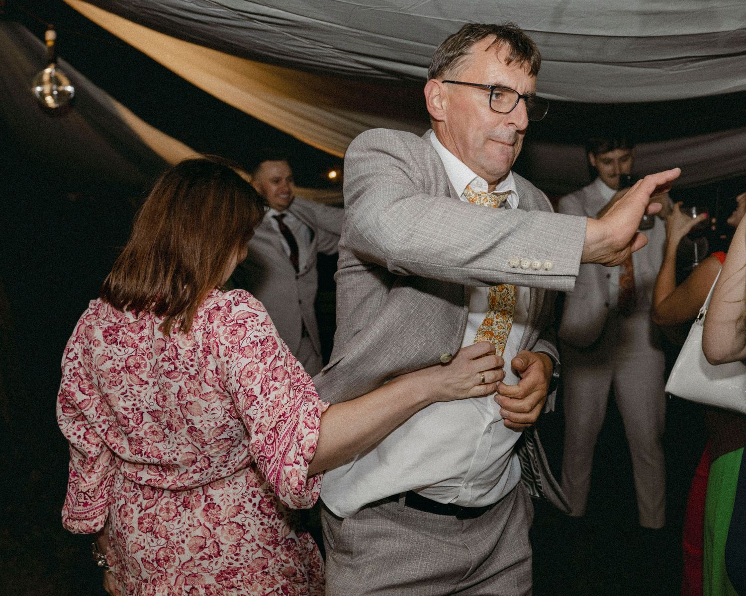 Elderly man in gray suit dancing energetically with woman in floral dress at a wedding reception.