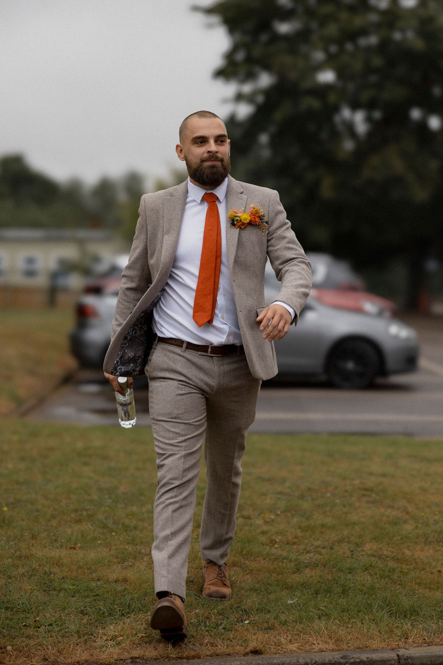 Bearded groom in beige suit with orange tie and boutonniere walks confidently across grass at outdoor wedding venue.