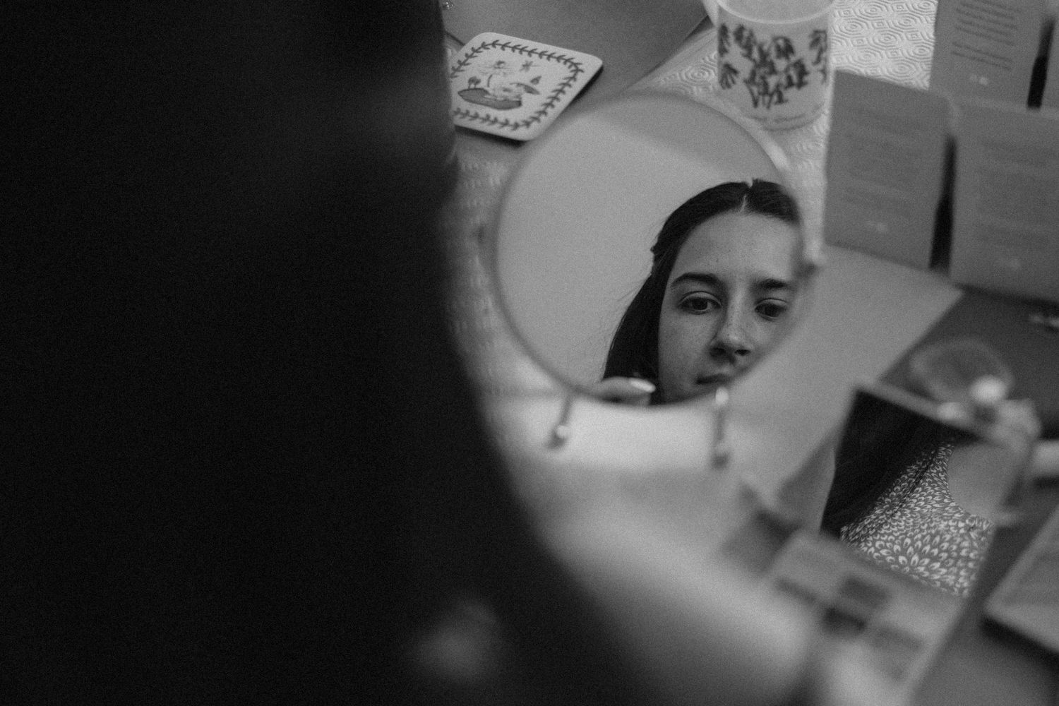 Black and white photo of a young woman looking at her reflection in a round vanity mirror on a dresser.