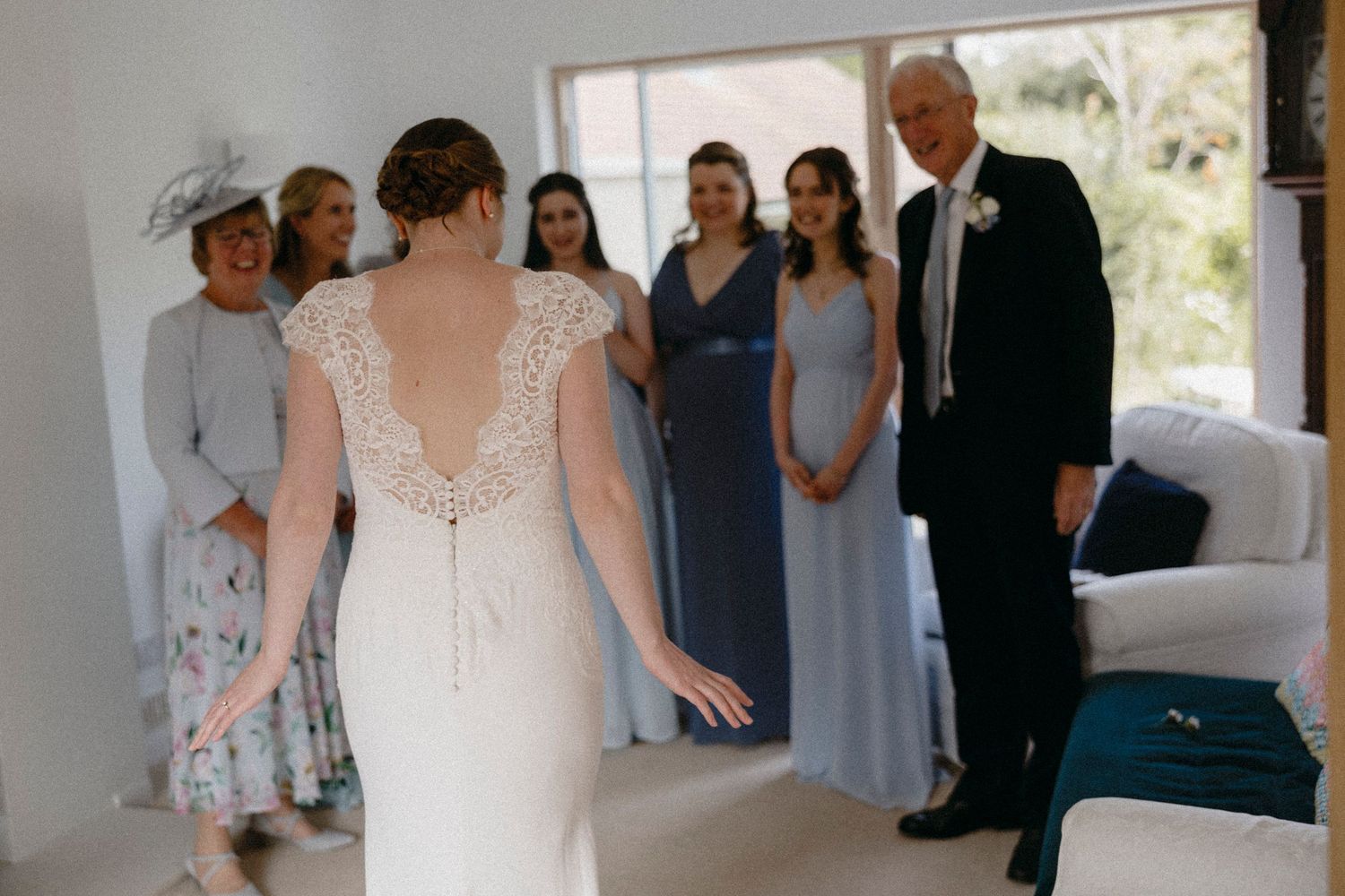 Bride in lace wedding dress reveals look to father and bridesmaids in blue gowns during bridal preparation.