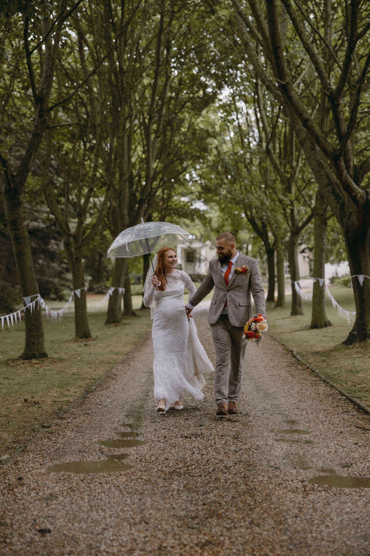 Bride and groom walk hand-in-hand down a tree-lined path in the rain, bride holding a clear umbrella.
