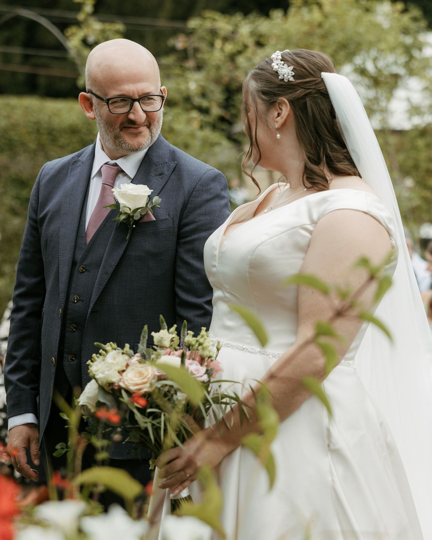 Groom in navy suit with glasses shares emotional moment with bride holding bouquet at outdoor wedding.