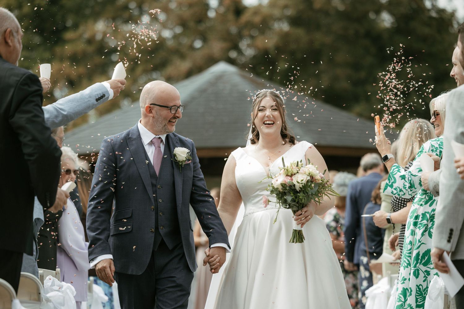Smiling bride and groom walking through confetti shower at outdoor wedding ceremony on sunny day.