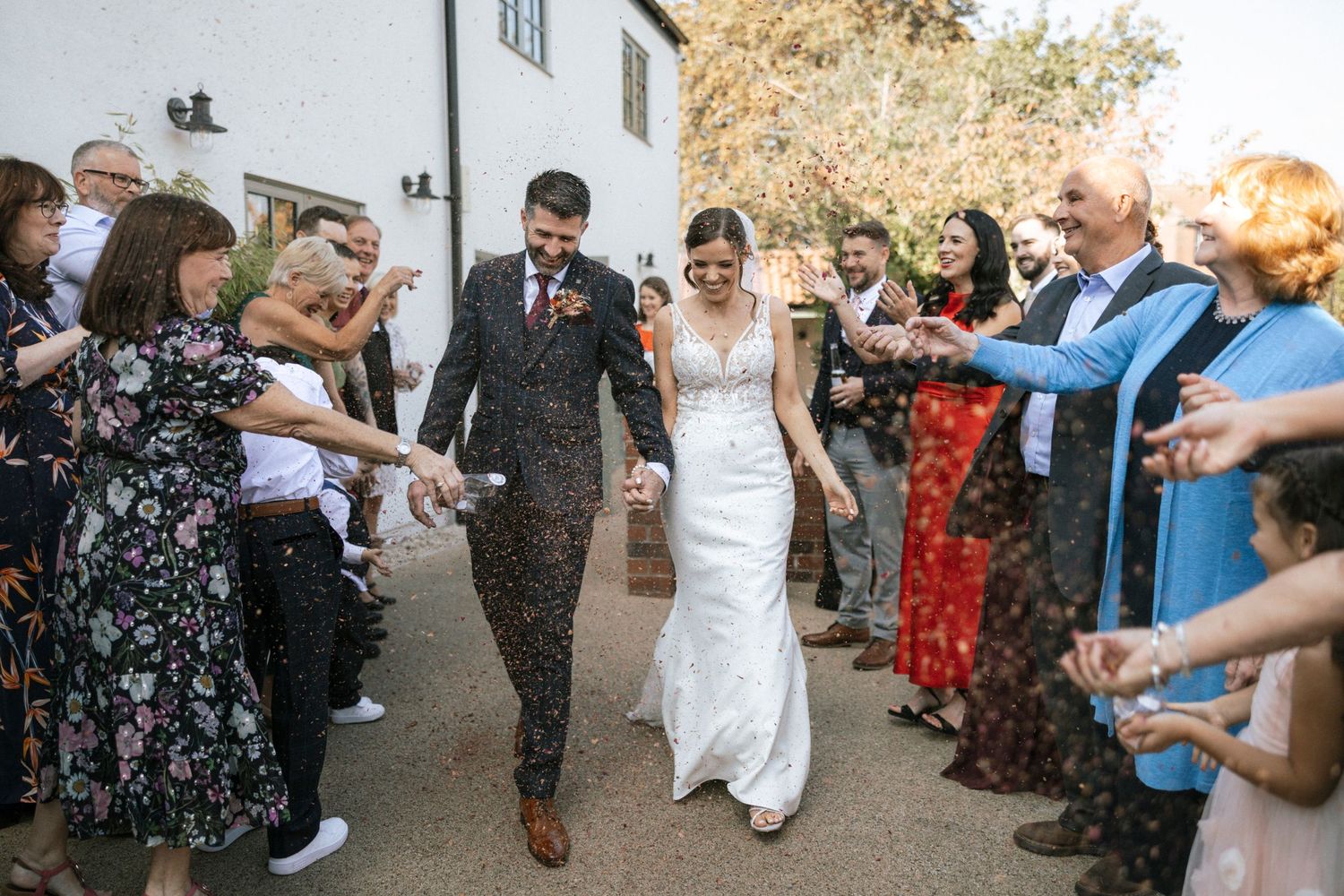 Bride and groom walk through confetti shower with smiling guests lining outdoor wedding venue path.