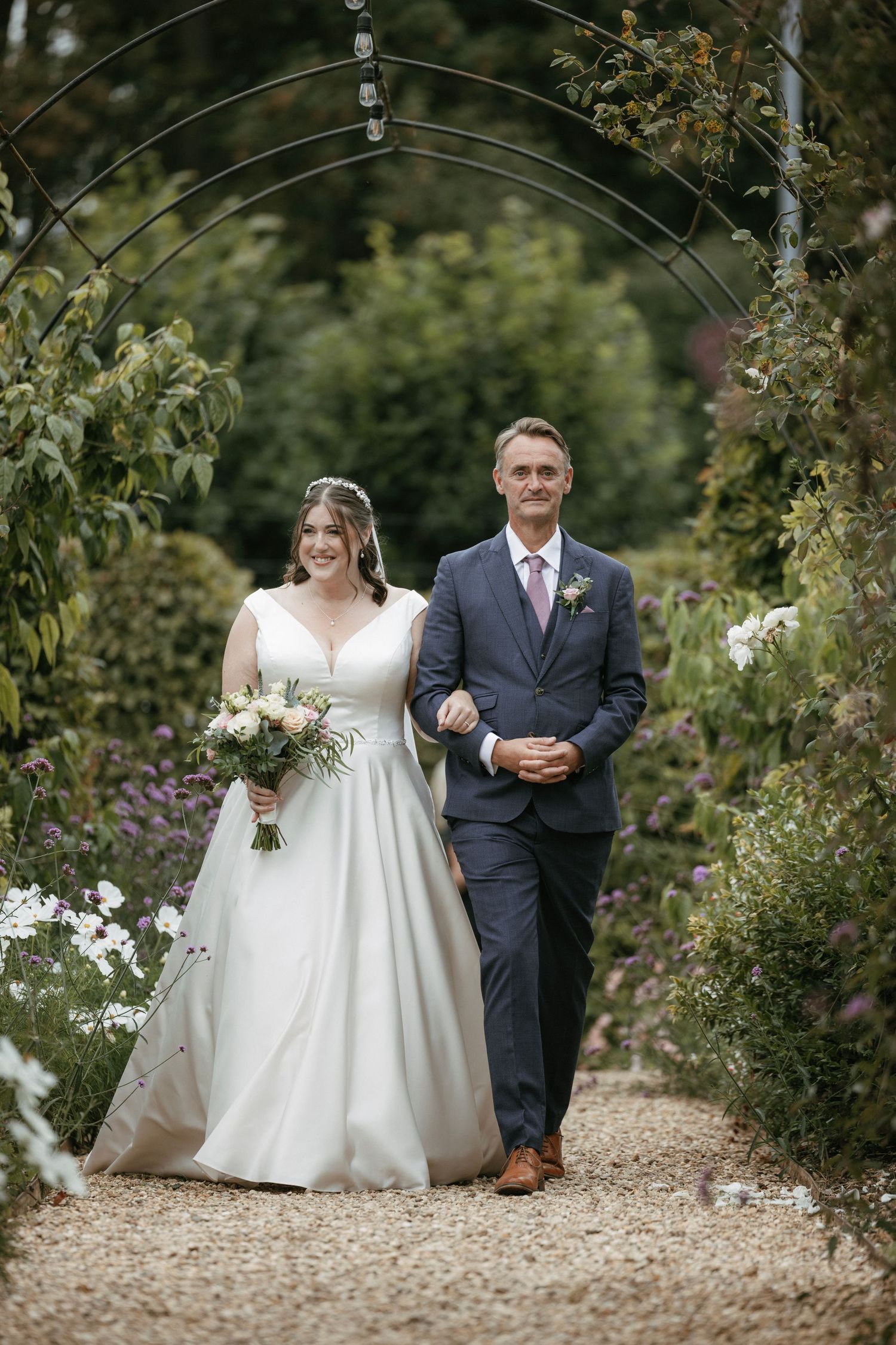 Bride in white gown walks with escort through garden arch on gravel path at outdoor wedding ceremony.