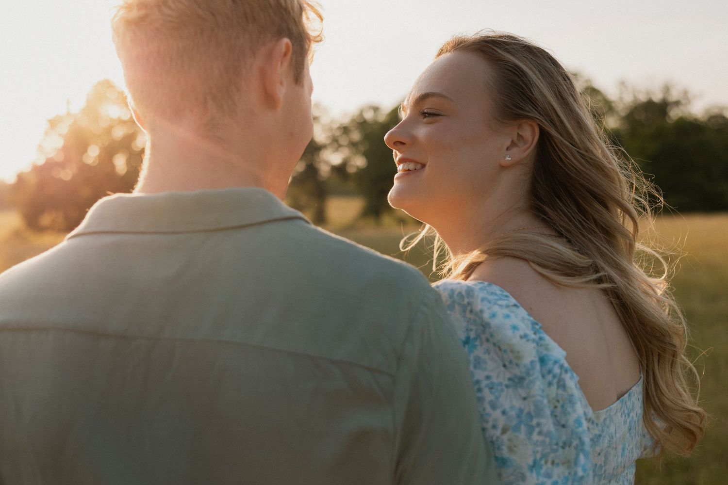 A smiling woman in a floral dress gazes at a man outdoors during golden hour sunset in a green field.