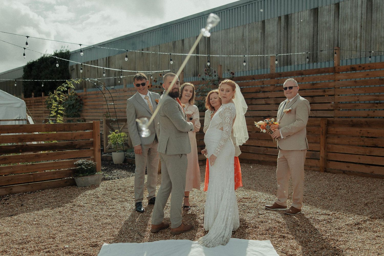 Wedding party taking a selfie with a selfie stick outdoors at a rustic barn venue with string lights.