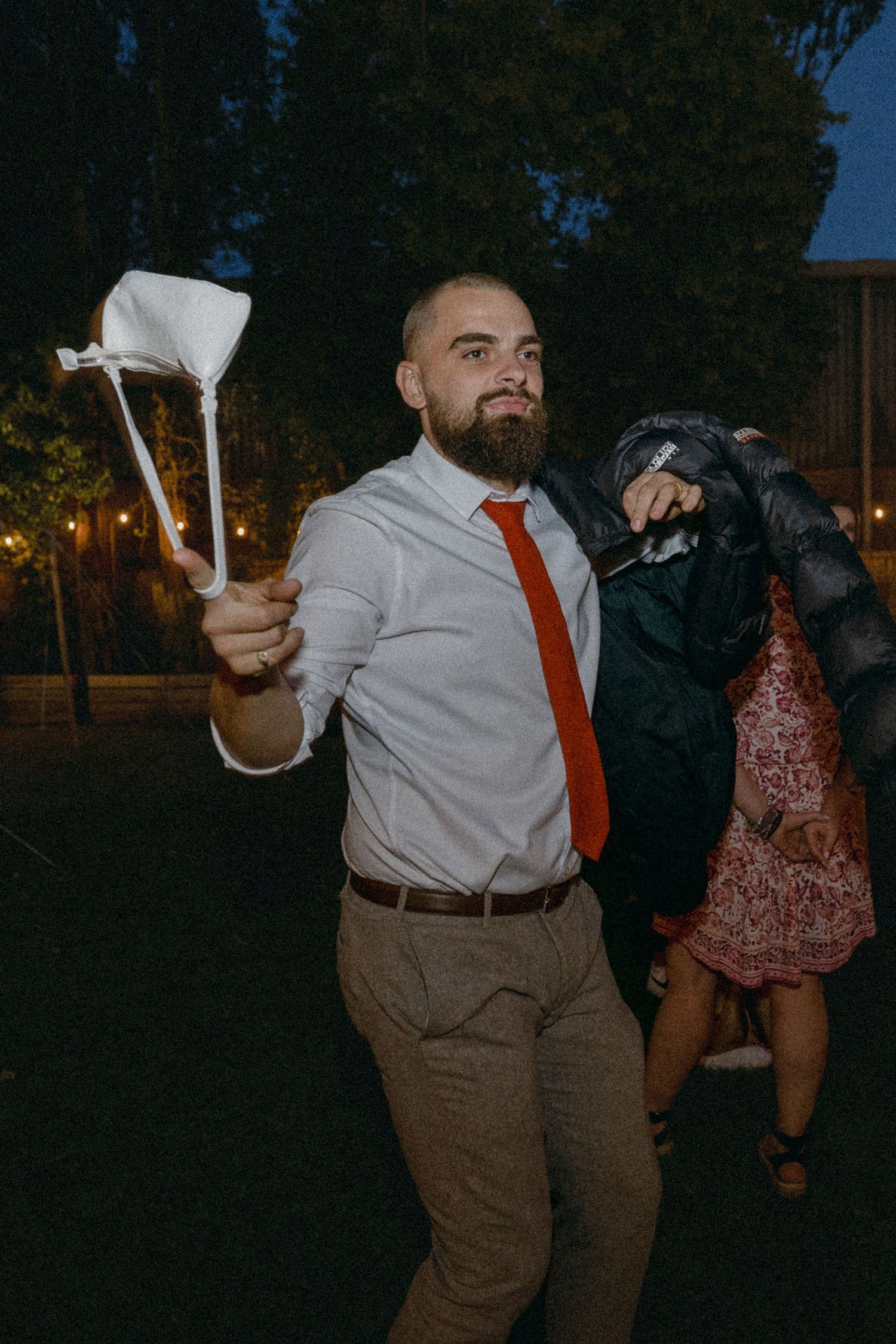 Bald bearded man in white shirt and red tie waving a white cloth at an outdoor evening event.