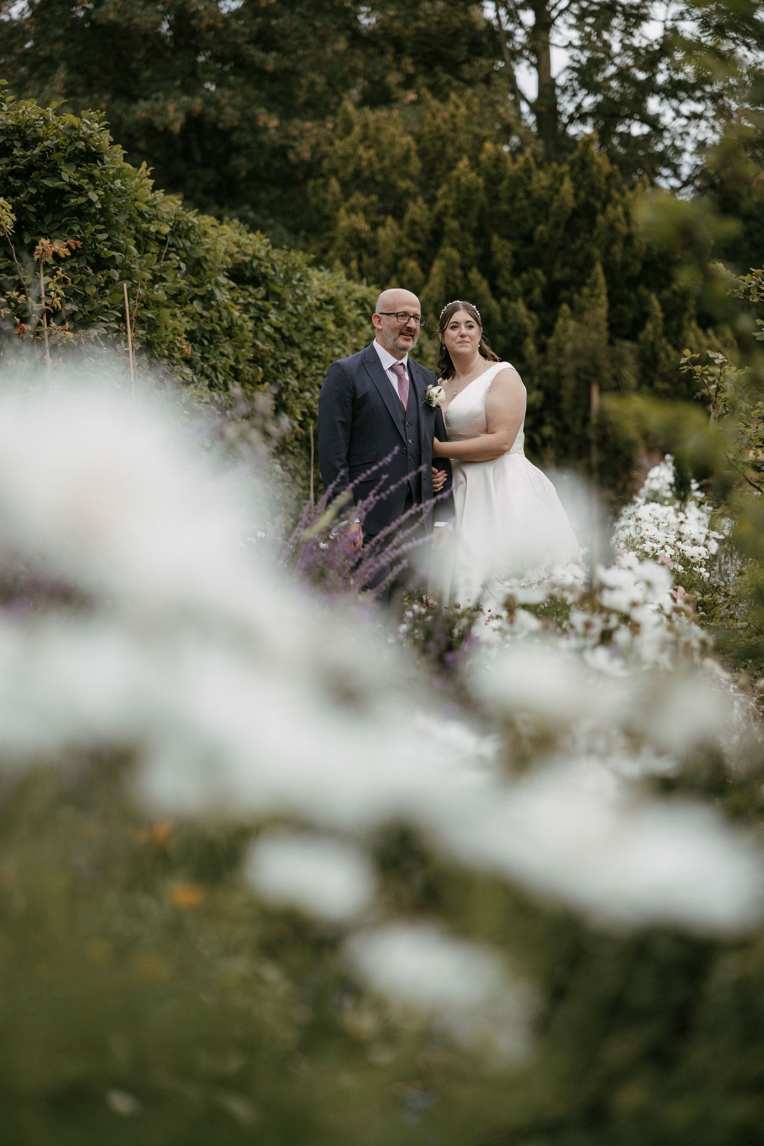 Bride and groom standing together in lush garden surrounded by lavender and white wildflowers.