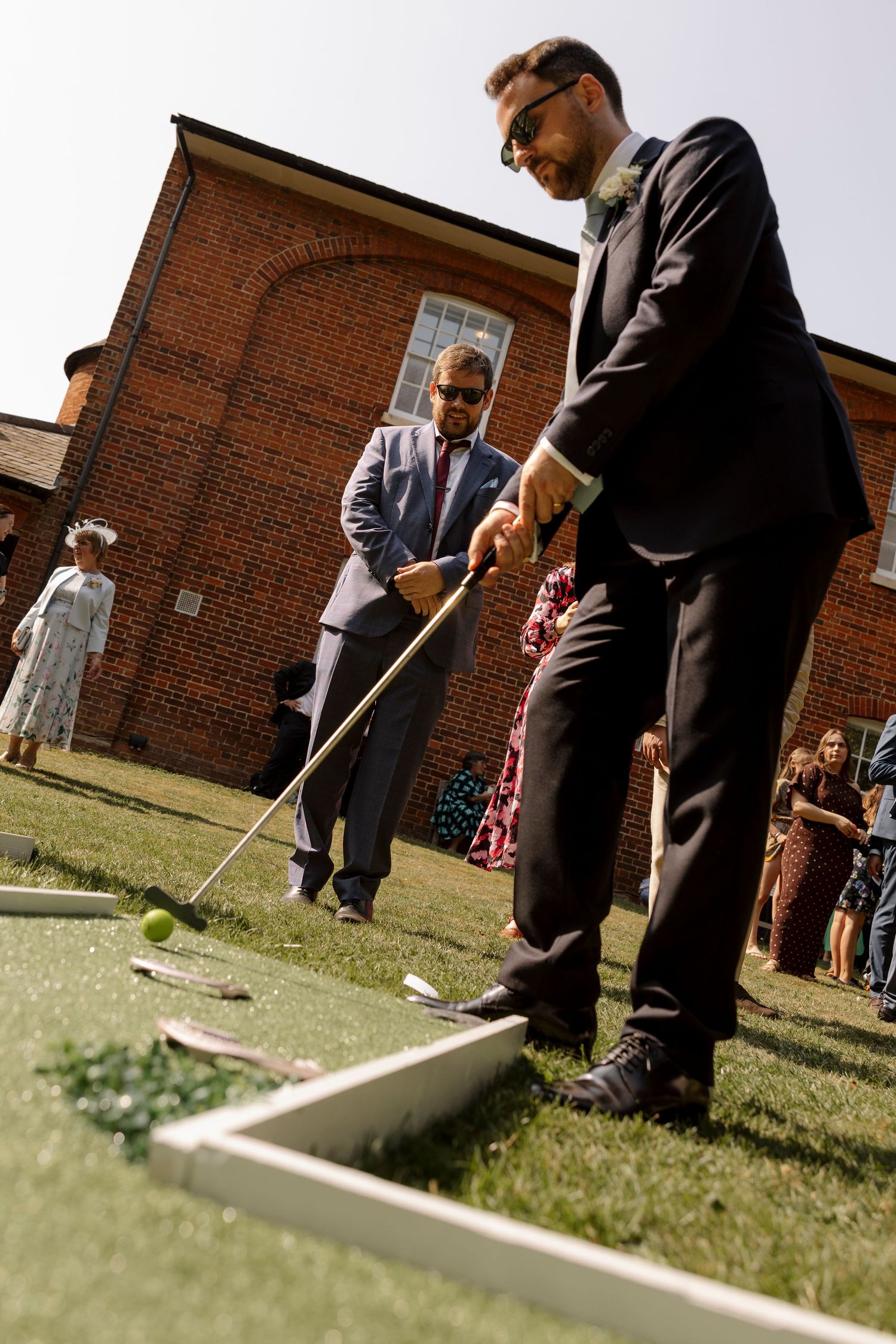 Two men in suits playing mini golf outdoors at a wedding reception near a brick building.