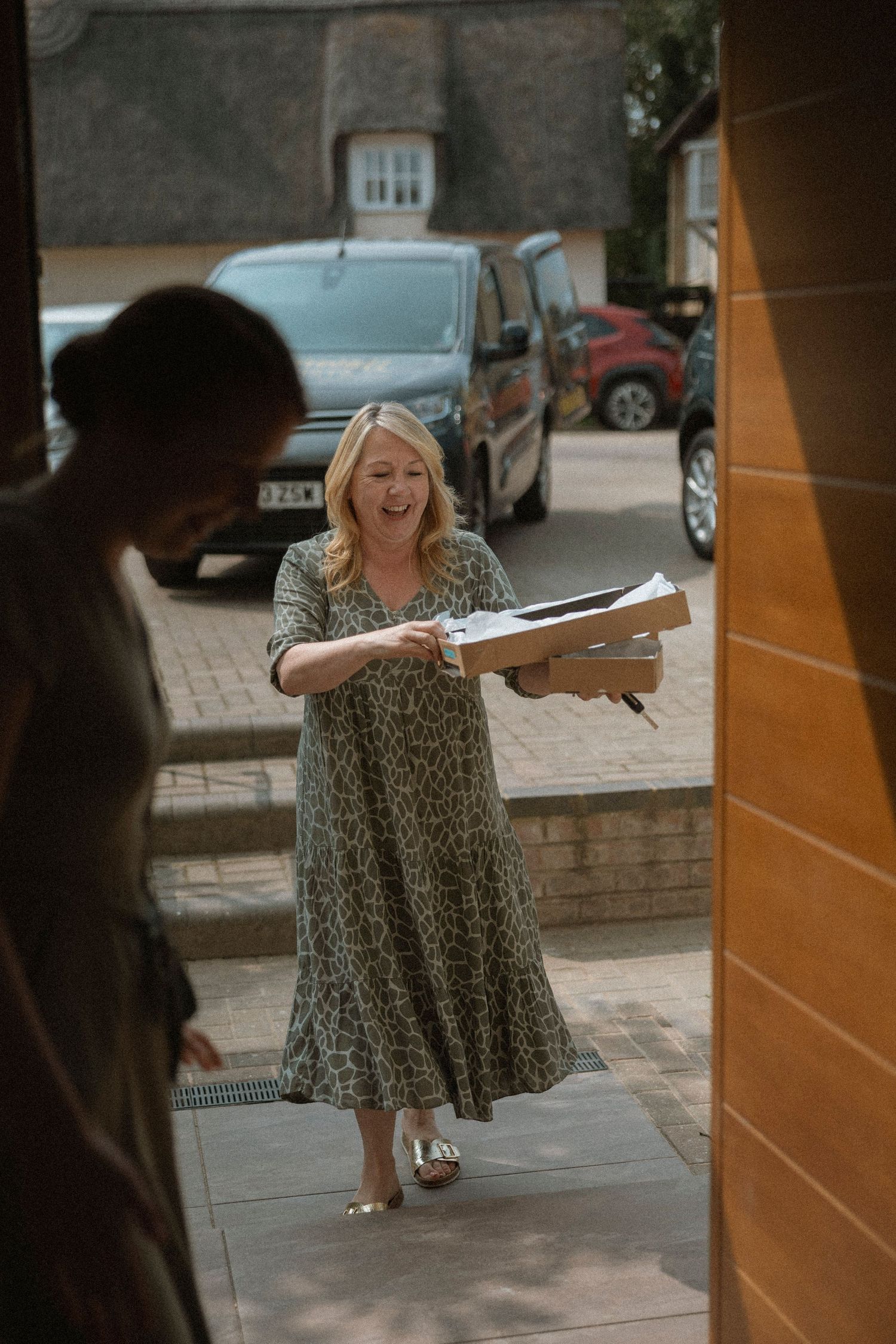 Smiling blonde woman in leopard print dress carrying boxes to a front door on a sunny day.