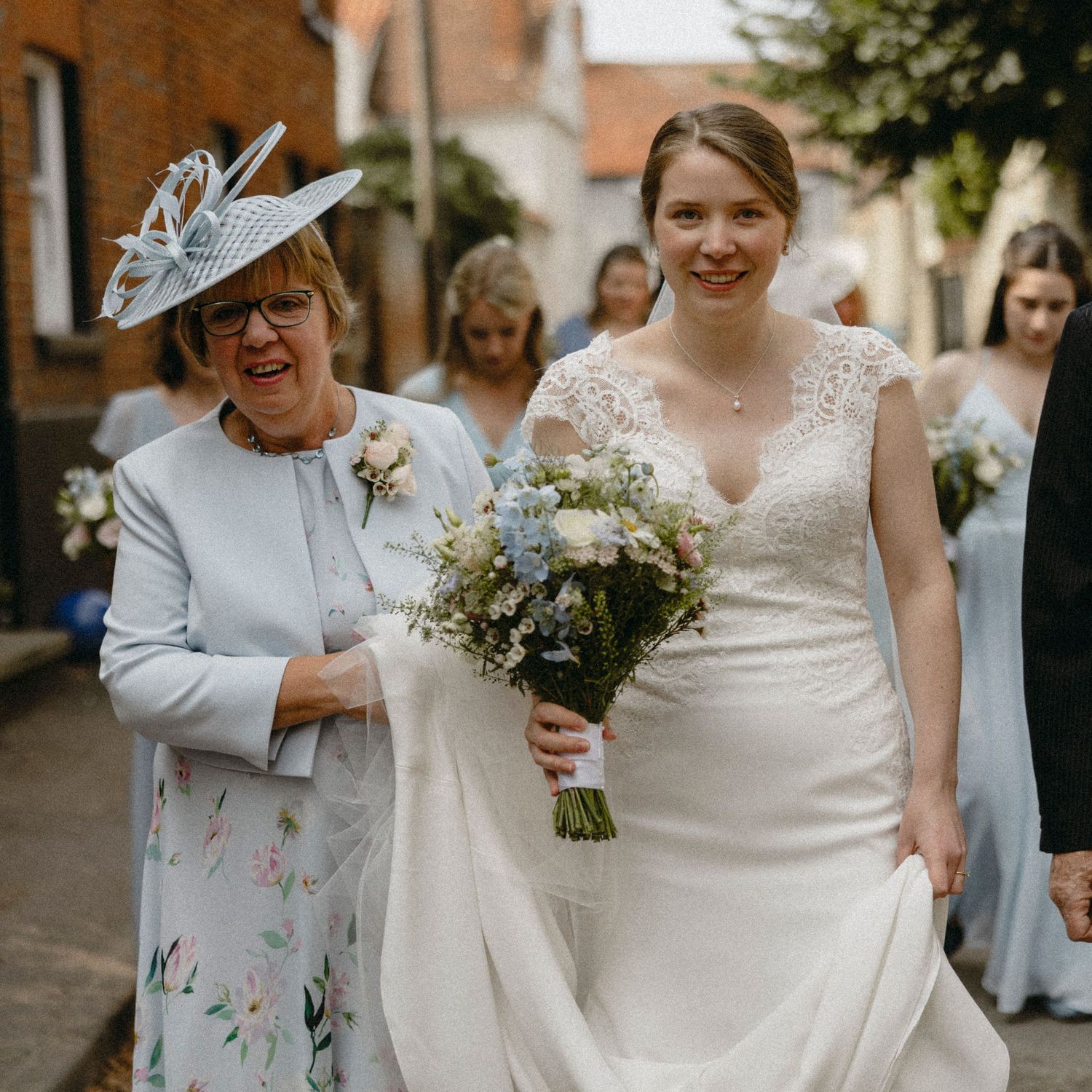 Bride in white lace gown walks with smiling older woman in blue hat, holding wildflower bouquet on cobblestone street.