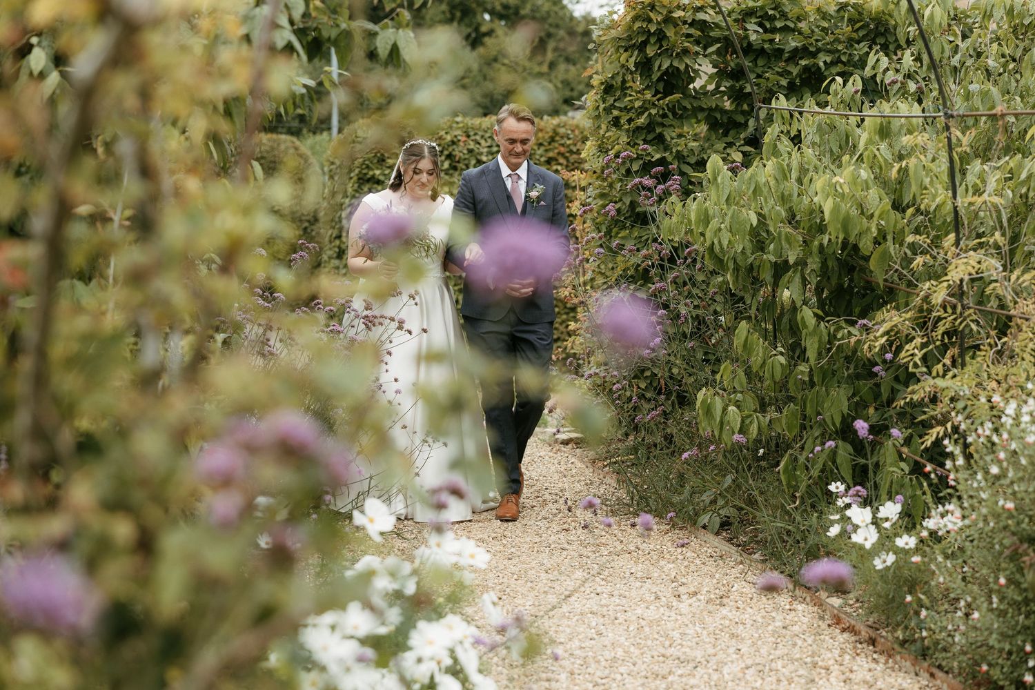 Bride and groom walking hand in hand along a garden path lined with purple flowers and lush greenery.