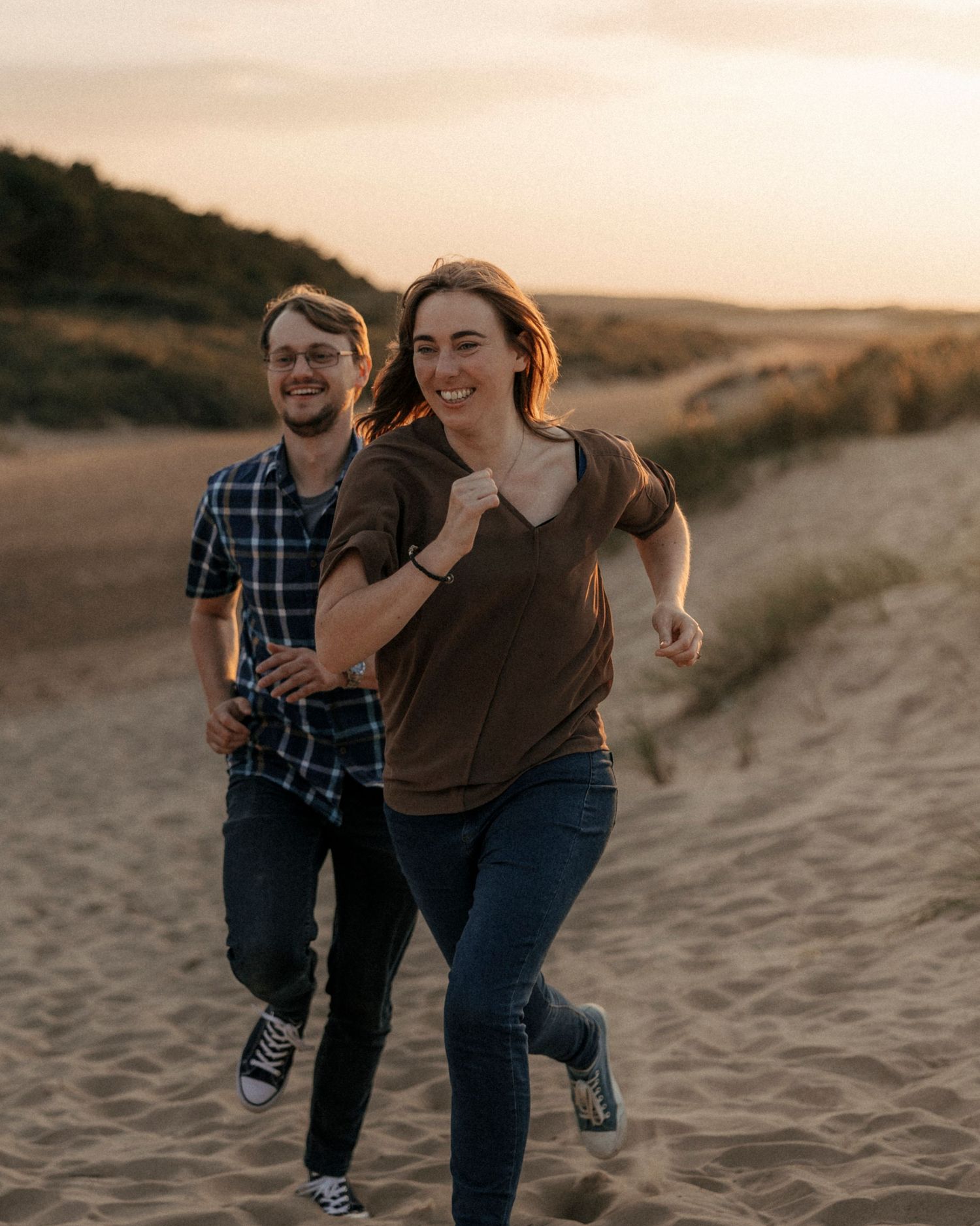 Happy couple running and laughing together on sandy dunes at golden hour sunset.