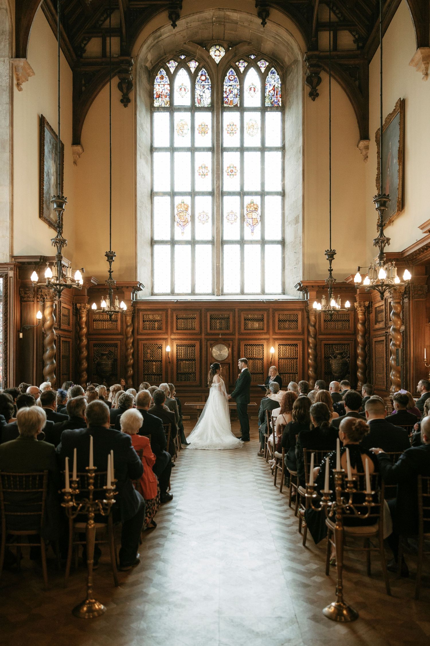 Elegant wedding ceremony in a grand hall with stained glass windows, wooden paneling, and candlelit aisle.