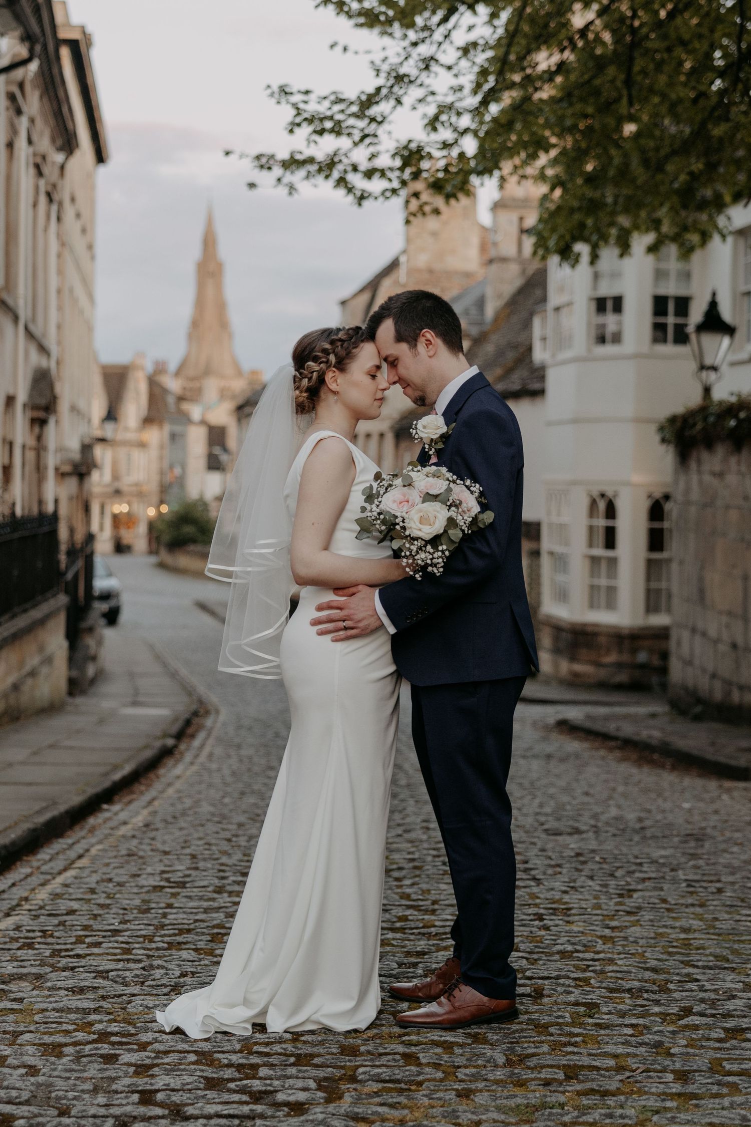 Bride and groom share romantic moment on cobblestone street at dusk, bride holding white bouquet.