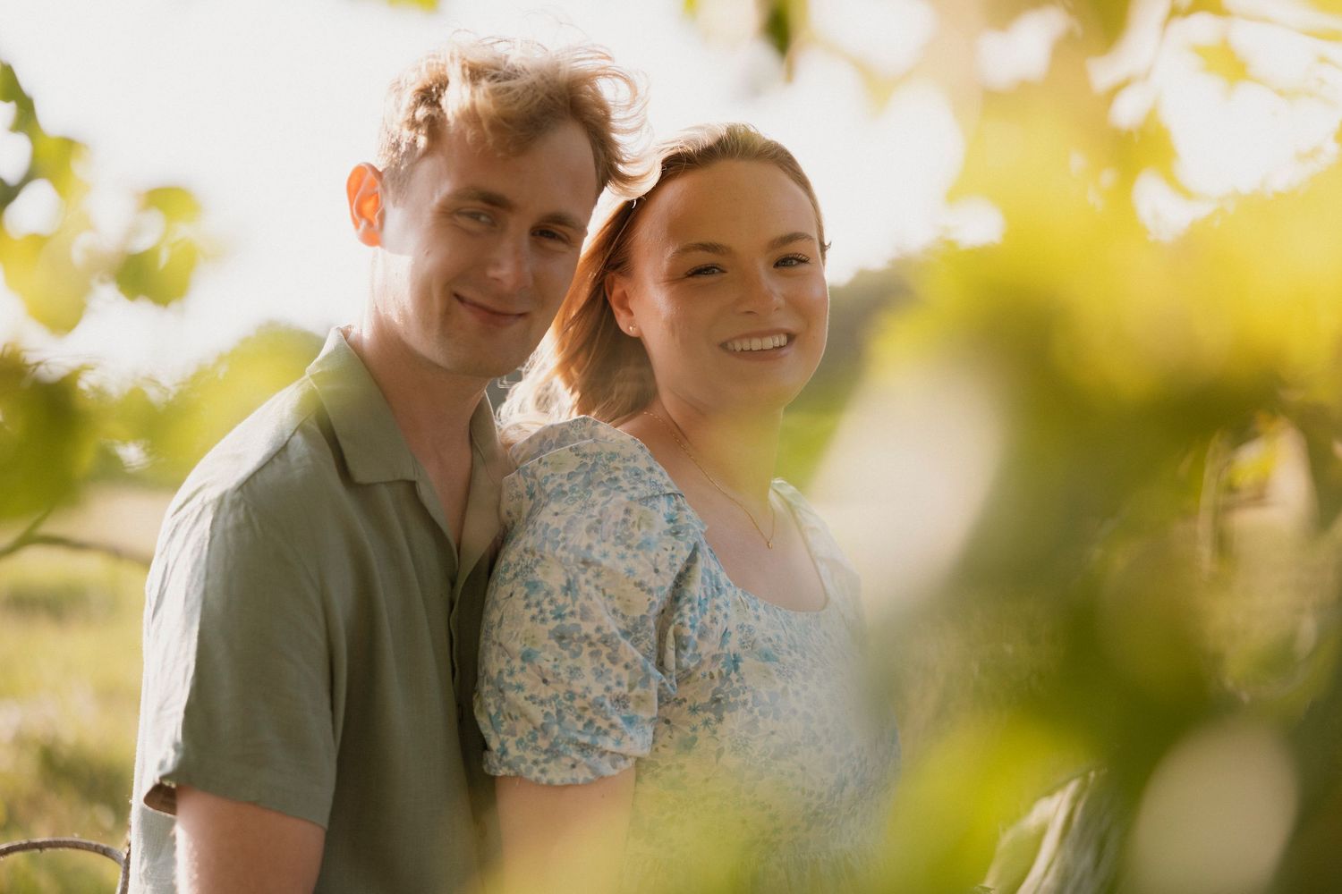 A couple smiles warmly among green vineyard leaves in golden sunlight.