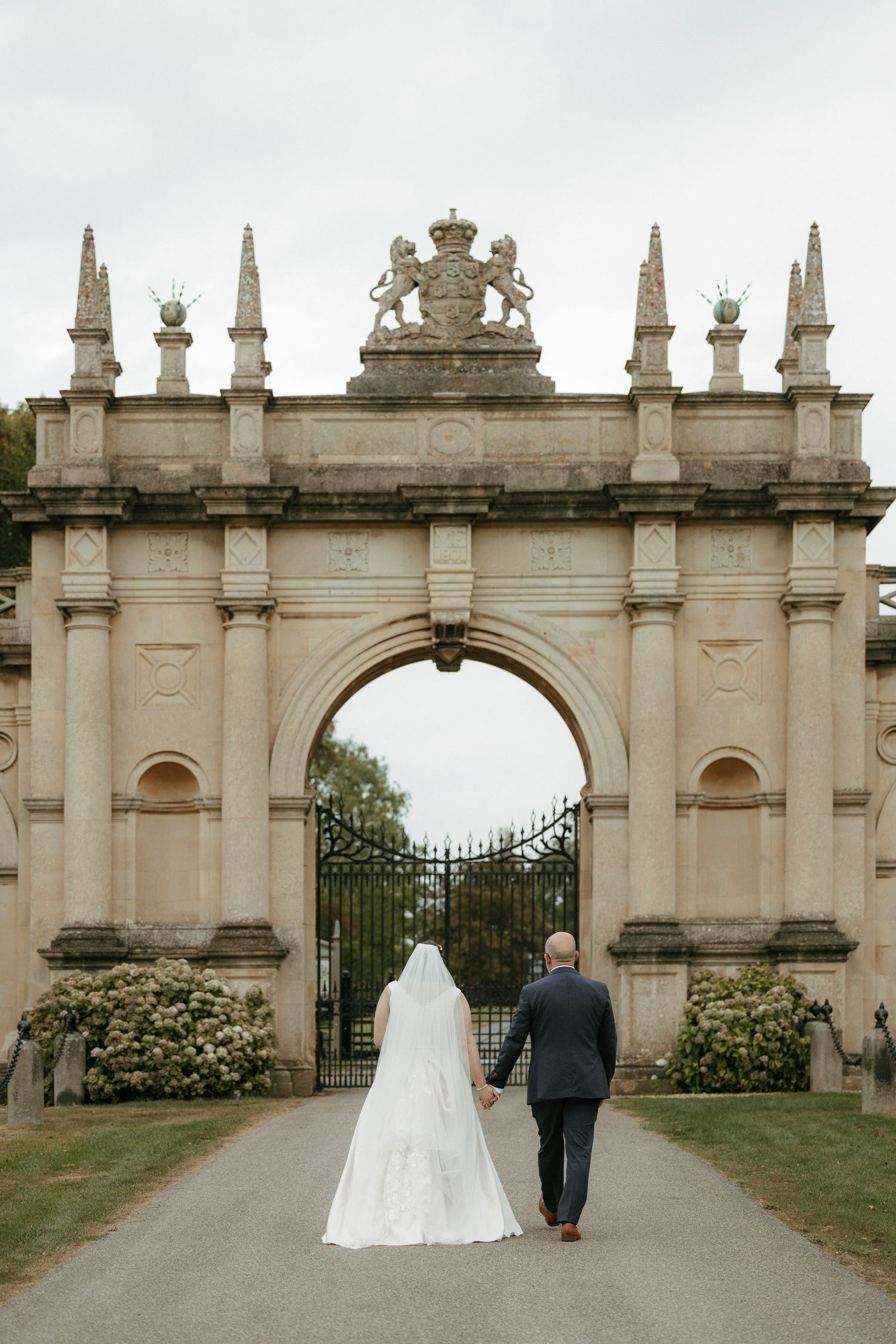 Bride and groom walking hand in hand toward an ornate stone arch gate at a grand estate wedding venue.