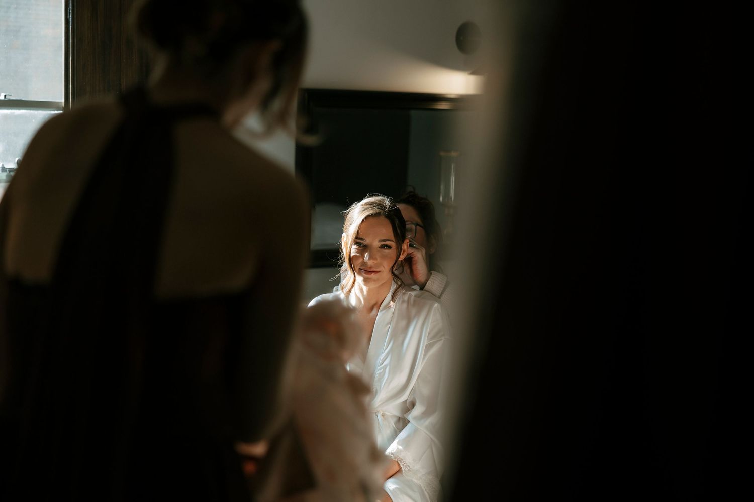 Bride getting ready in soft light, assisted by bridesmaid in black dress, captured in moody wedding preparation photo.