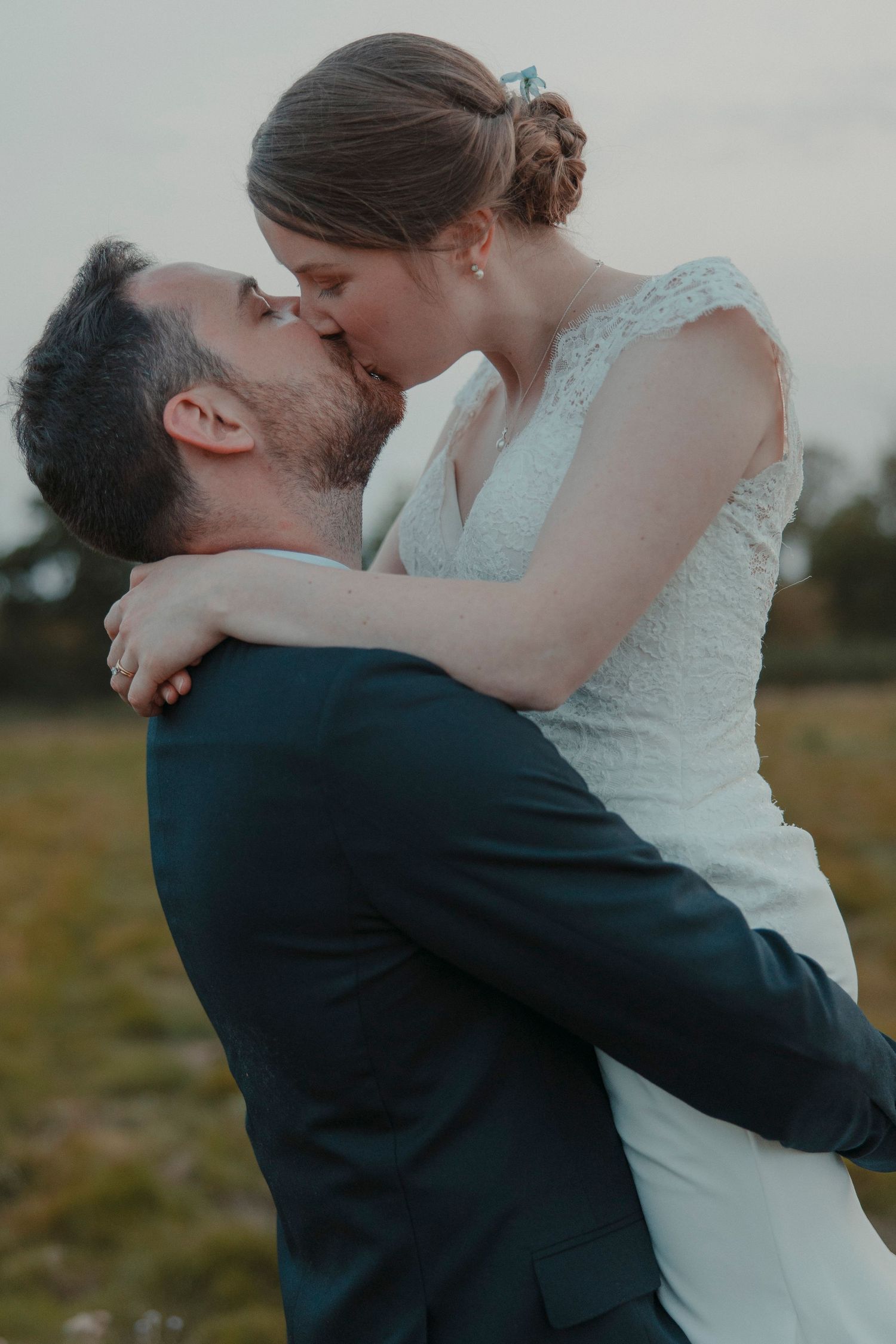 A groom in a dark suit lifts and kisses his bride in a white lace wedding dress outdoors.