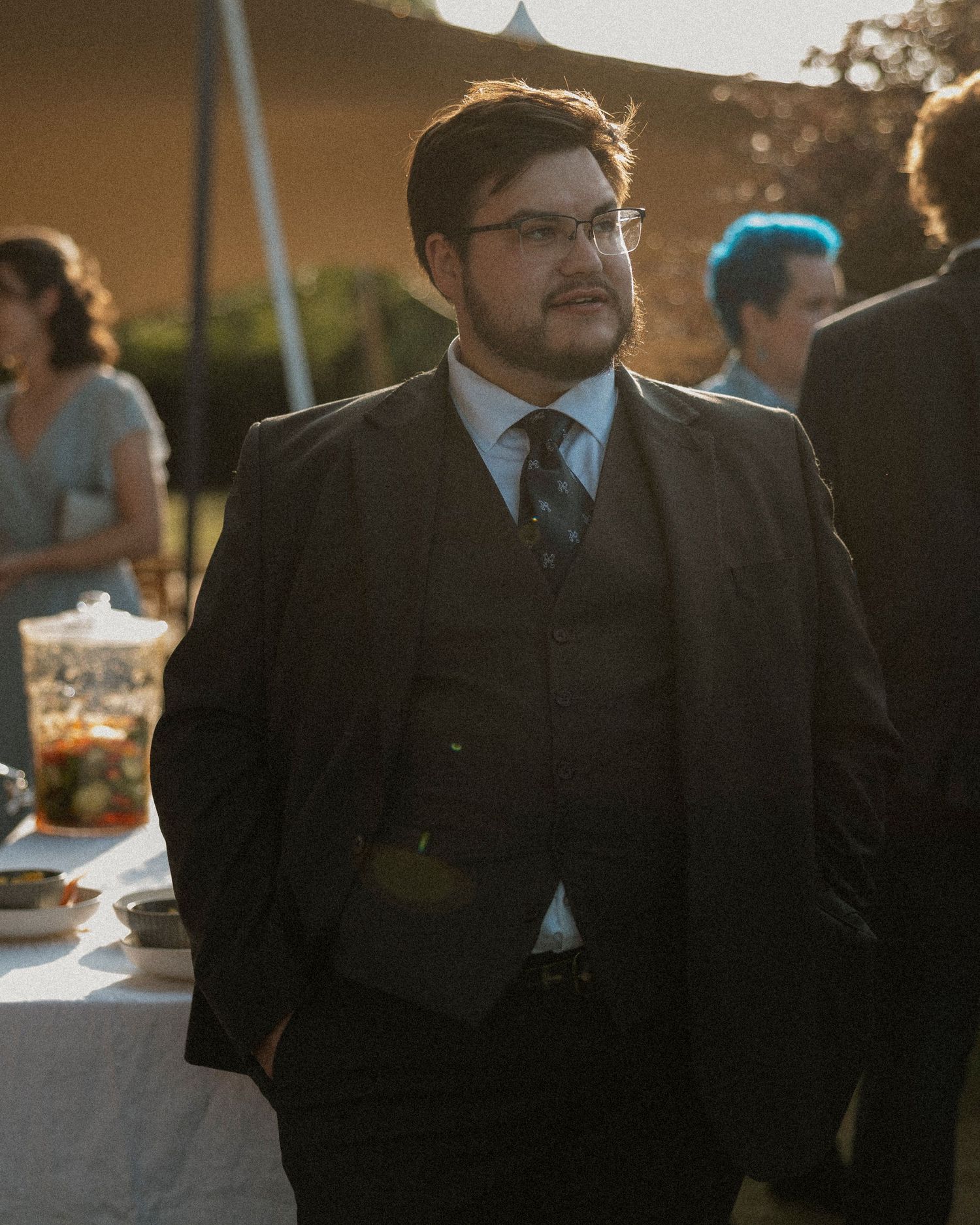 Young man in dark suit and tie with glasses at outdoor evening event.