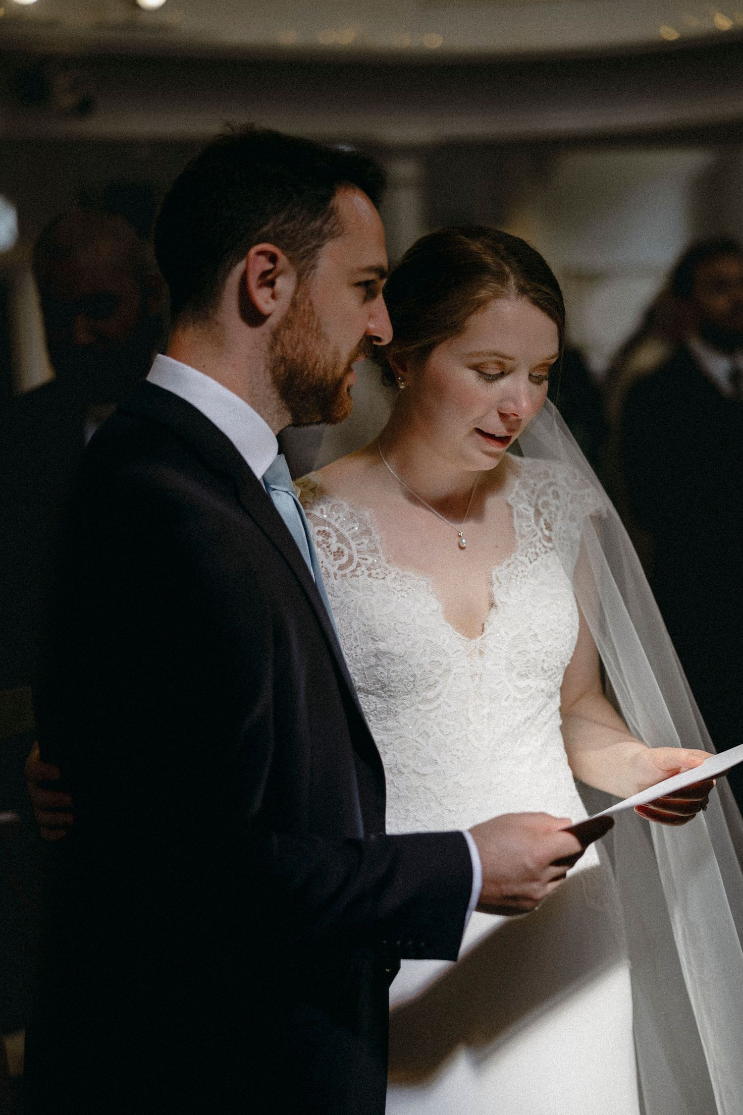 Bride and groom reading wedding vows together during ceremony, bride in lace dress with veil, groom in dark suit.