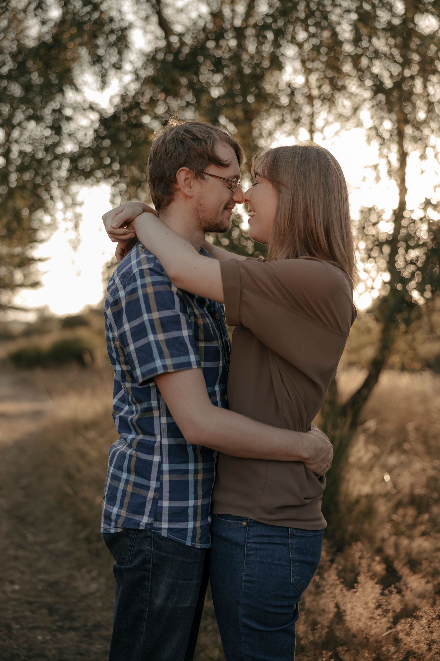 A couple embraces and smiles at each other outdoors at golden hour near a tree.