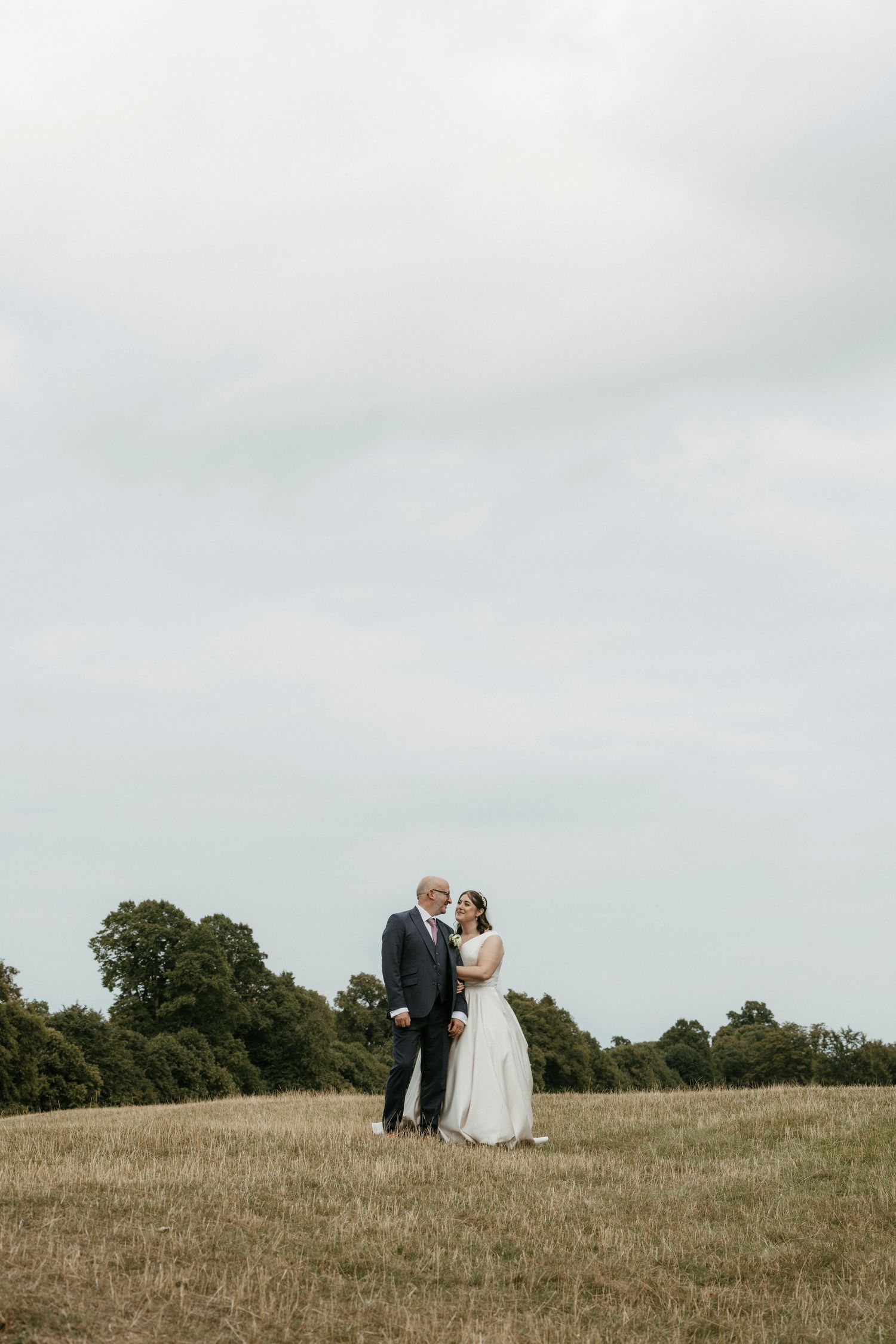 A bride and groom stand together in an open field with trees and overcast sky on their wedding day.