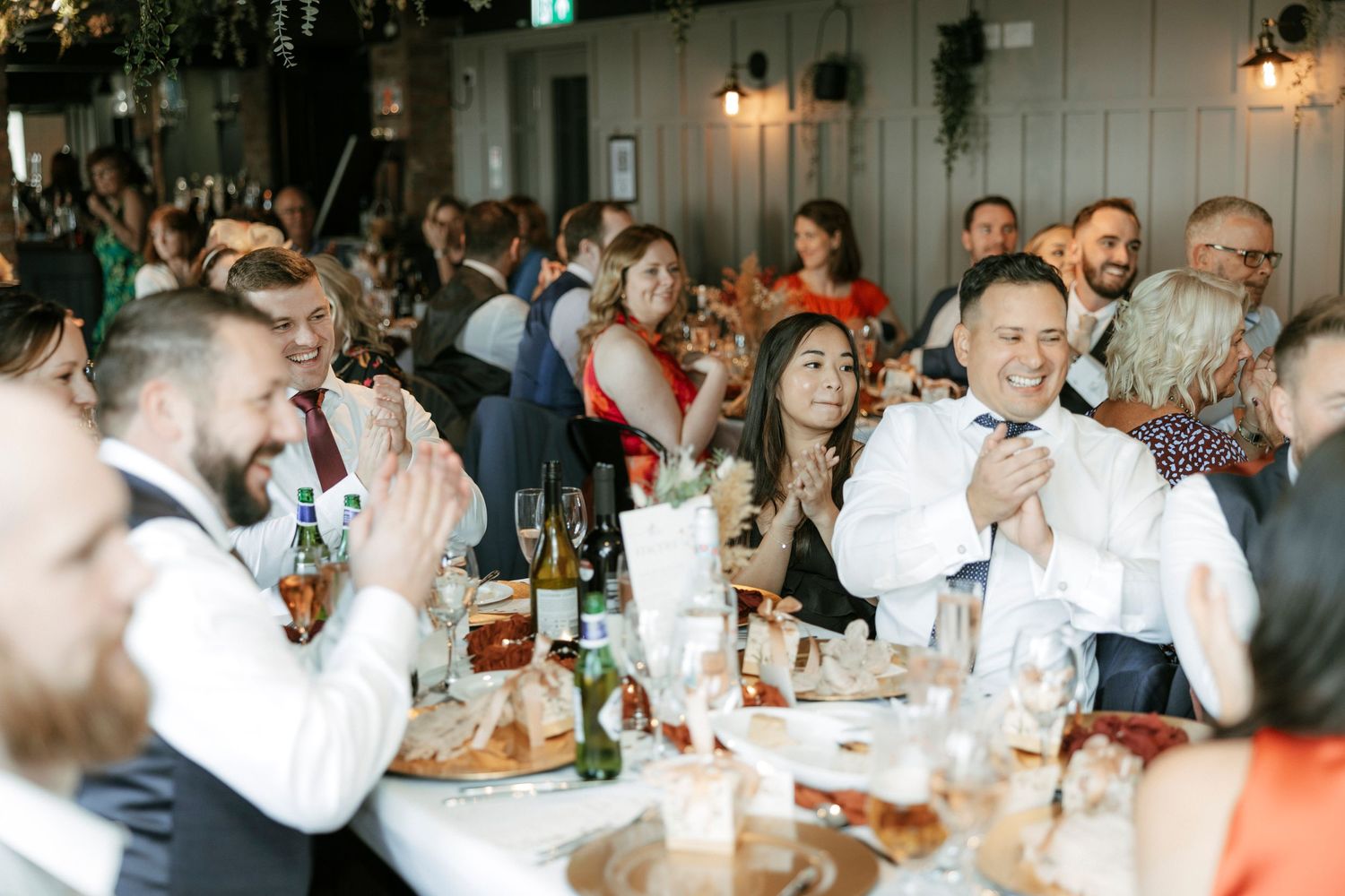 Wedding reception guests laugh and enjoy themselves at a long dining table filled with drinks and table settings.