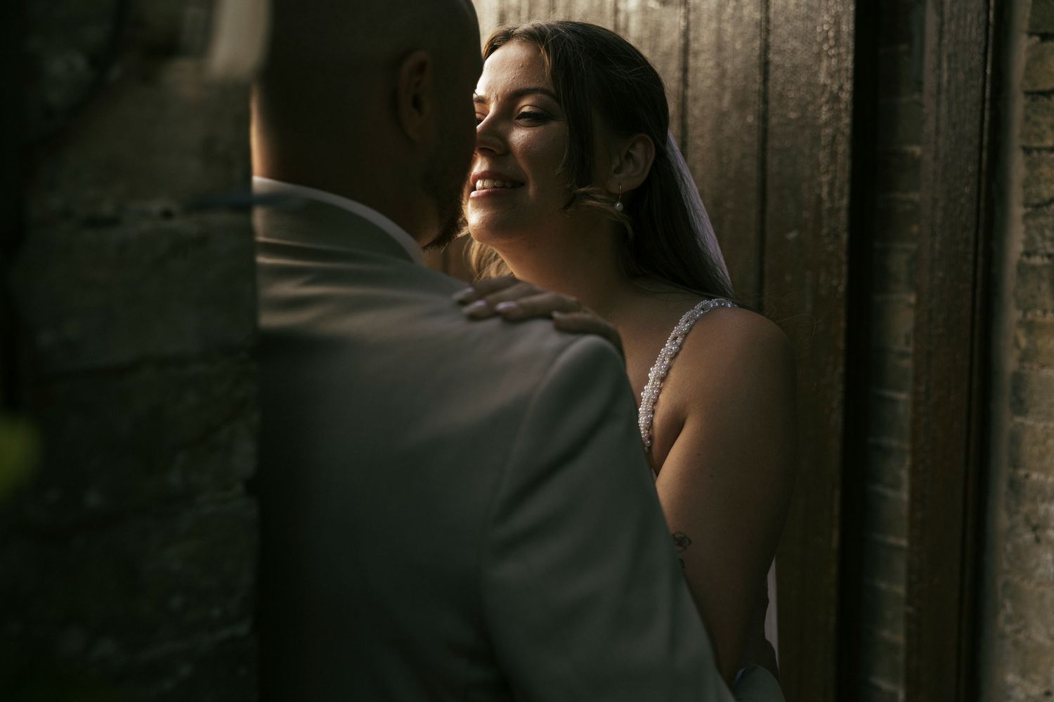 A bride in a white dress stands in dramatic side lighting near a window, creating an intimate wedding portrait.