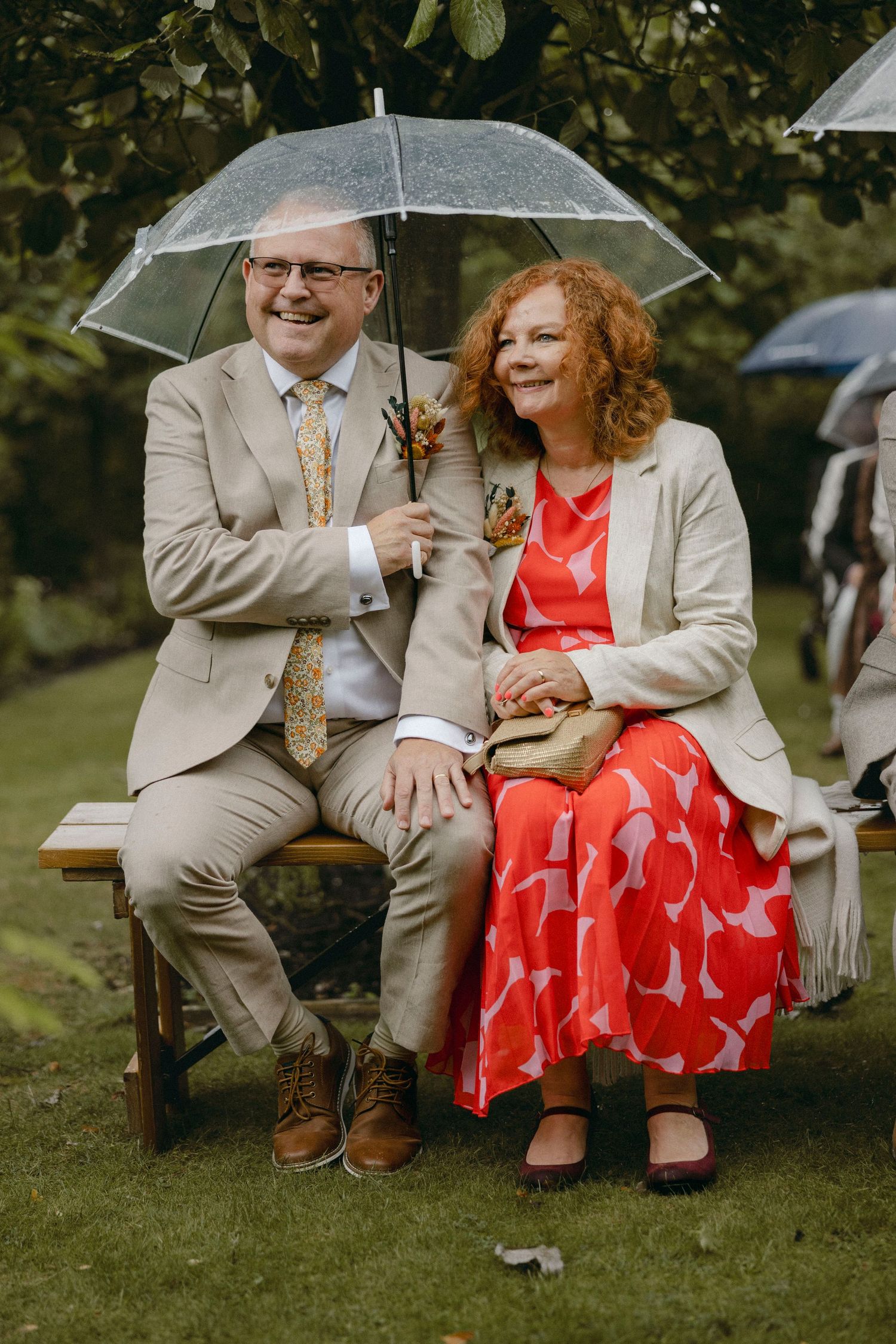 Smiling couple sitting on bench sharing clear umbrella outdoors, man in tan suit with floral tie, woman in red patterned dress.