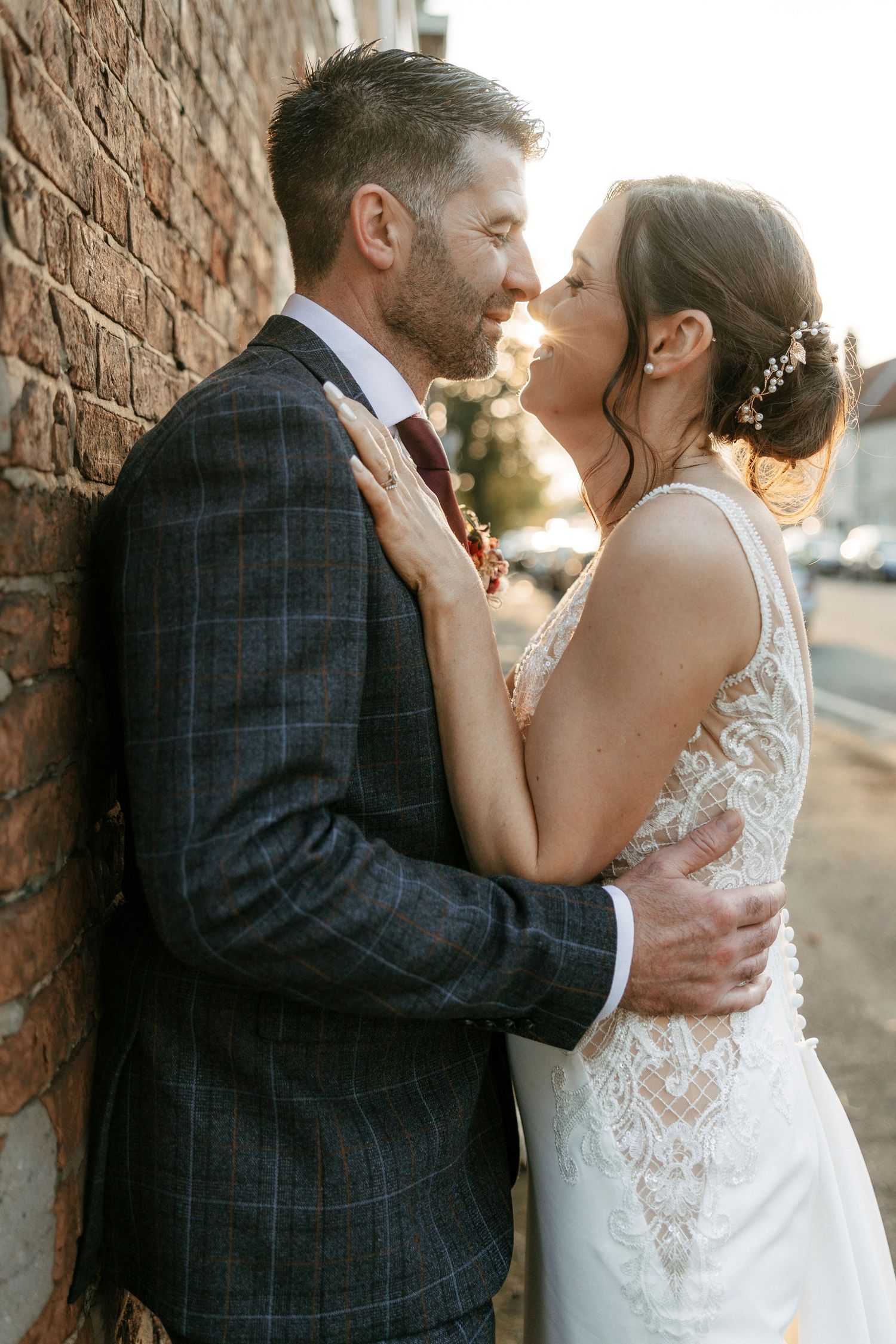 Newlyweds share intimate moment against brick wall at sunset.
