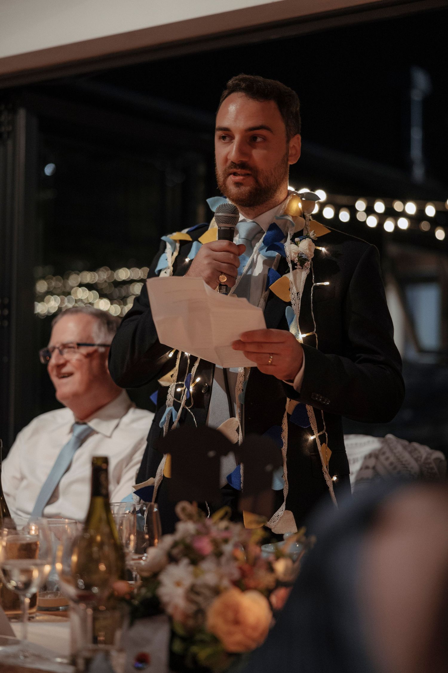 A man in a suit delivers a wedding speech holding a microphone and papers, adorned with colorful ribbons at a reception dinner.